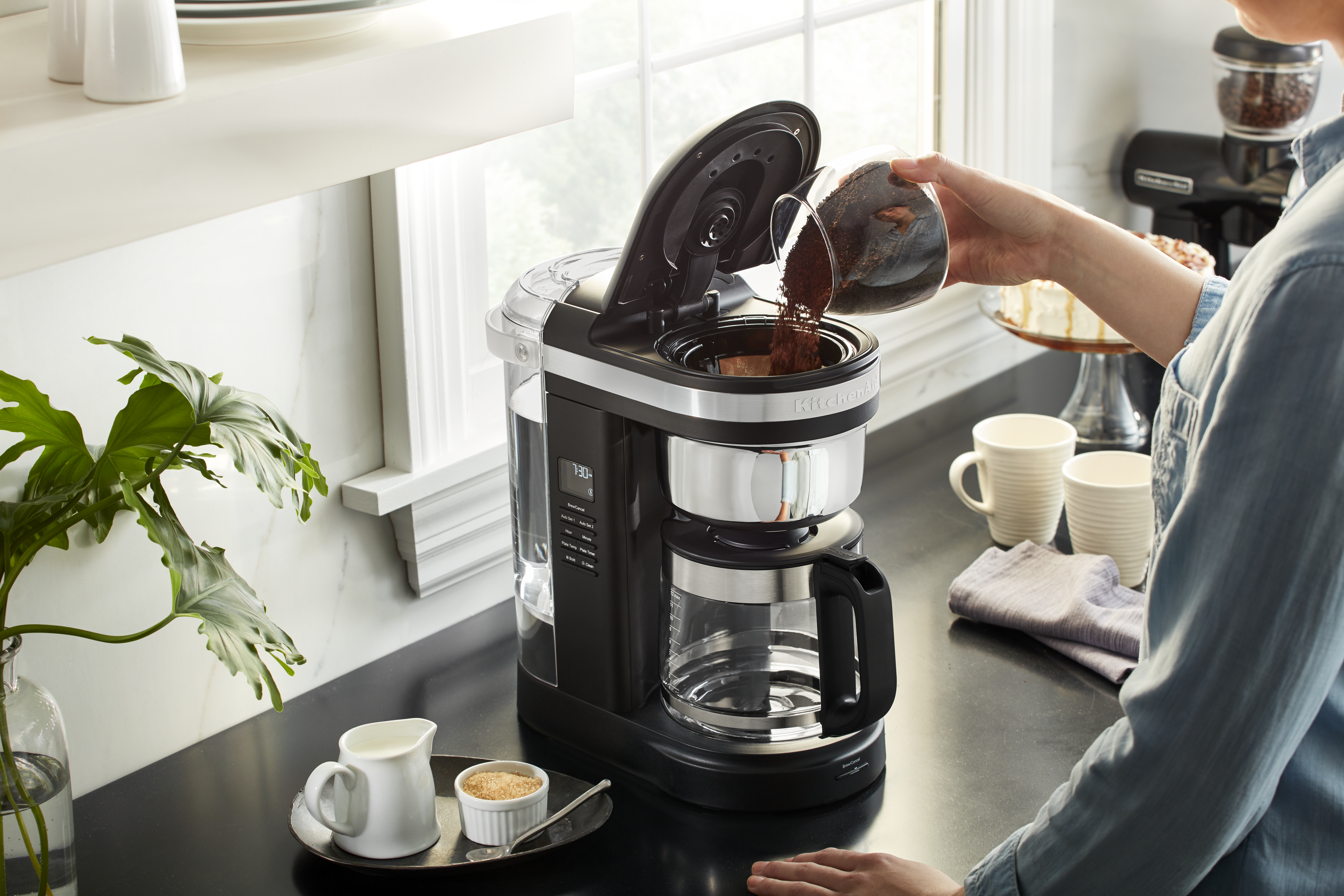 Eine Person gießt Kaffeepulver in eine schwarze Kaffeemaschine. Auf der Theke befinden sich eine Pflanze, Zucker, Kaffeeweißer und Tassen. Im Hintergrund ist ein Kuchen.