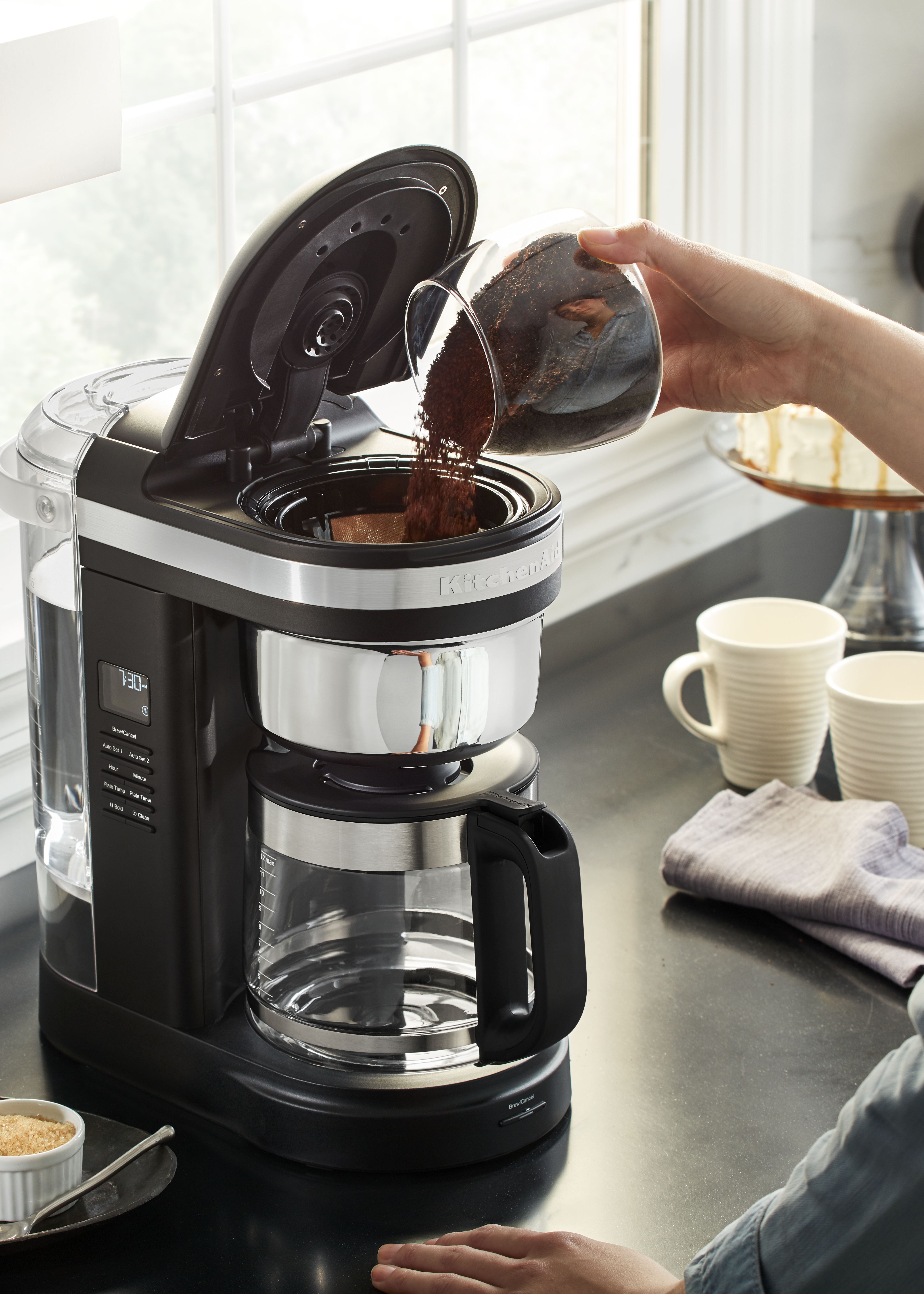 Eine Kaffeemaschine mit einer Person, die Kaffeepulver hineingießt. Der Hintergrund ist eine Küche.