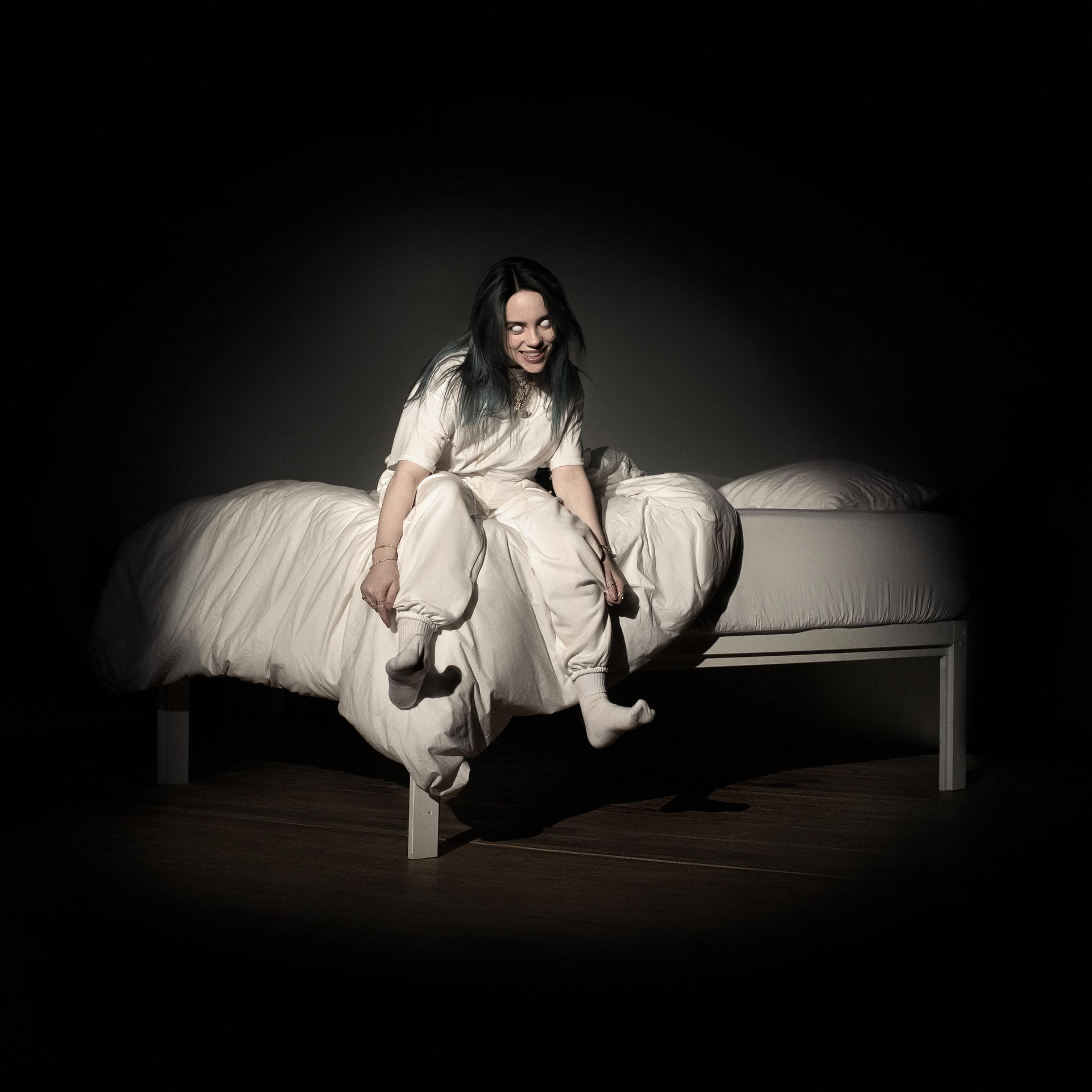Billie Eilish assise sur un lit, riant. Elle porte des vêtements blancs, fond sombre et éclairage vif.