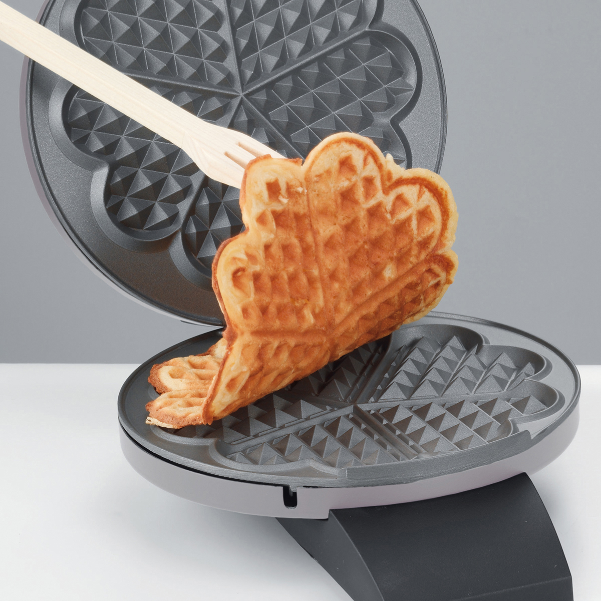 Waffeleisen mit einer Waffel, die mit einer Holzgabel entnommen wird. Die Waffel ist herzförmig.