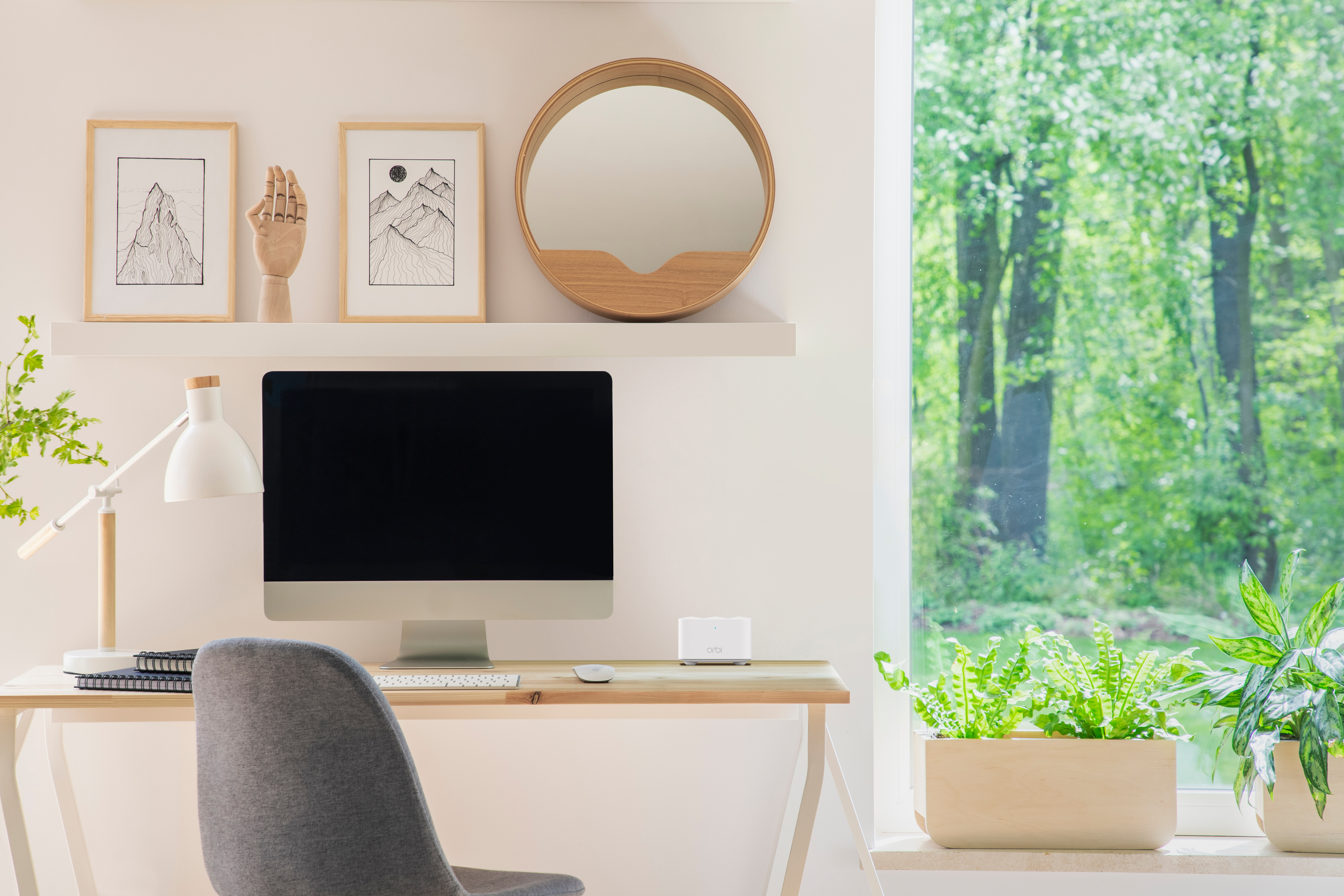 Schreibtisch mit Computer, Kunst und Blick auf Bäume. Ein grauer Stuhl steht vor dem Schreibtisch.