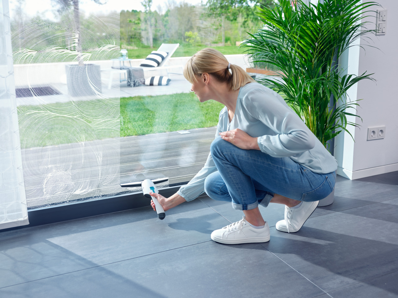 Frau putzt Fenster mit einem Abzieher. Sie hockt auf einem grauen Fliesenboden in der Nähe einer Pflanze.