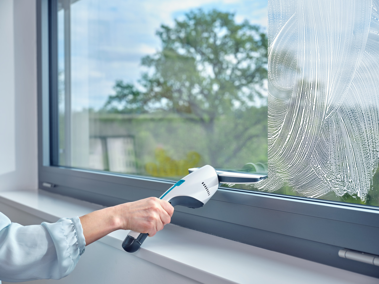 Eine Hand reinigt ein Fenster mit einem Reinigungswerkzeug. Draußen sind ein grüner Baum und blauer Himmel.