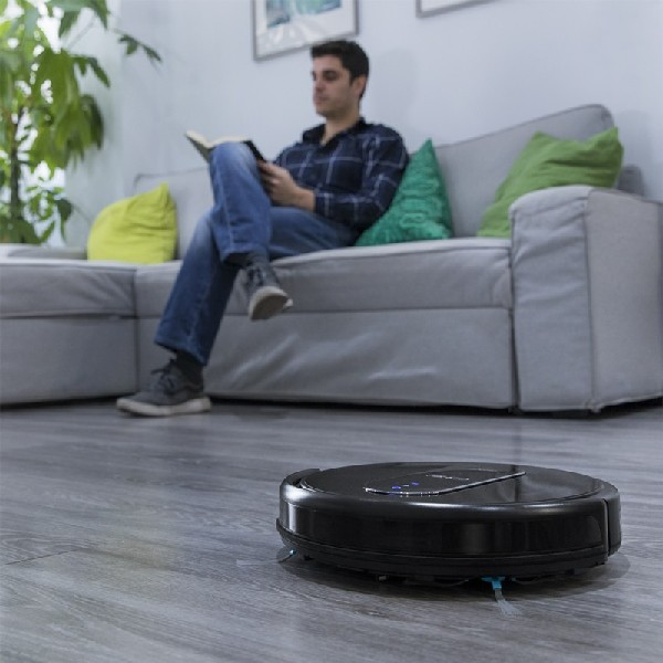 Aspiradora robótica en piso de madera, hombre leyendo en sofá gris en el fondo.