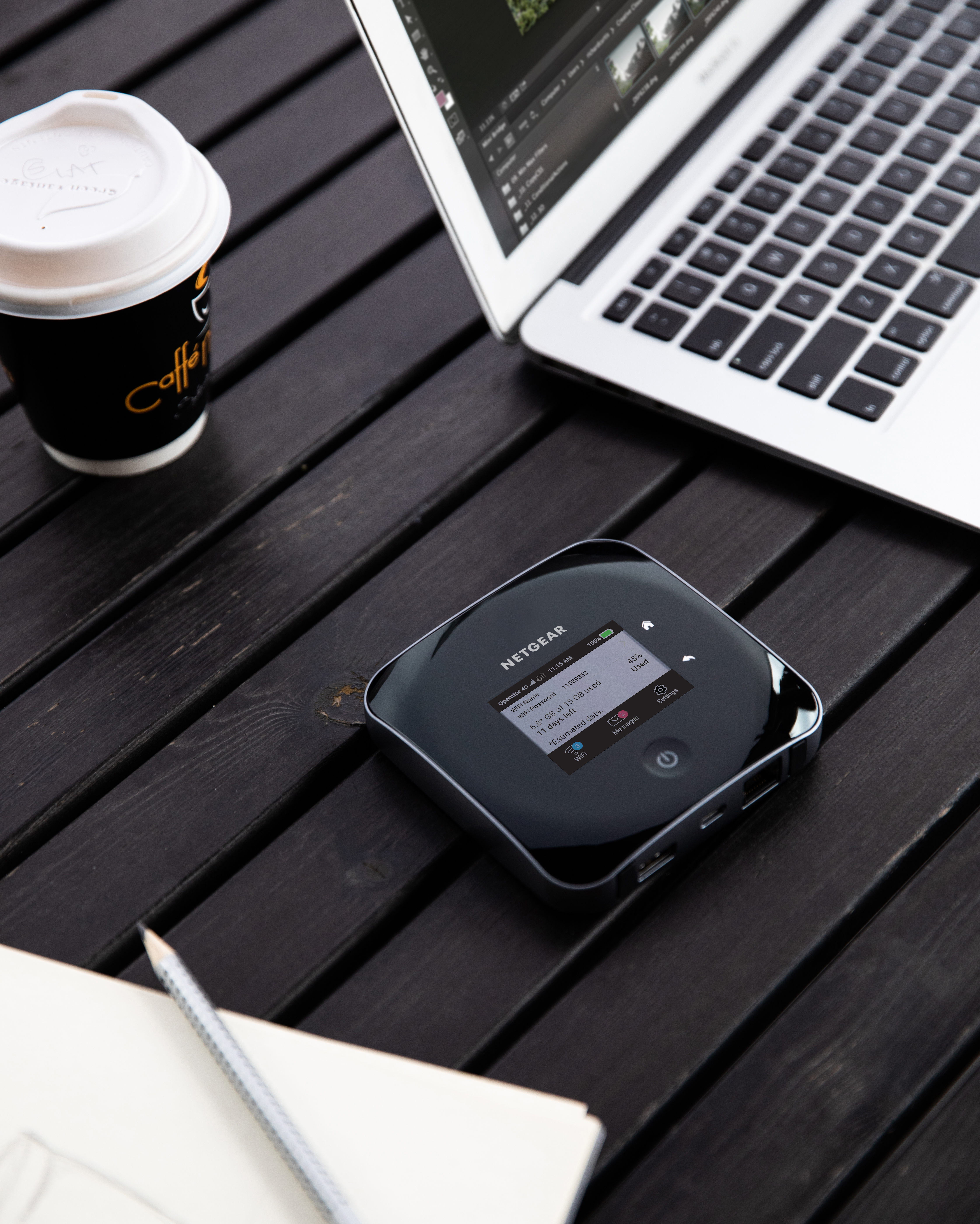 A black wireless router, laptop, coffee cup, and pencil sit on a dark wooden table.