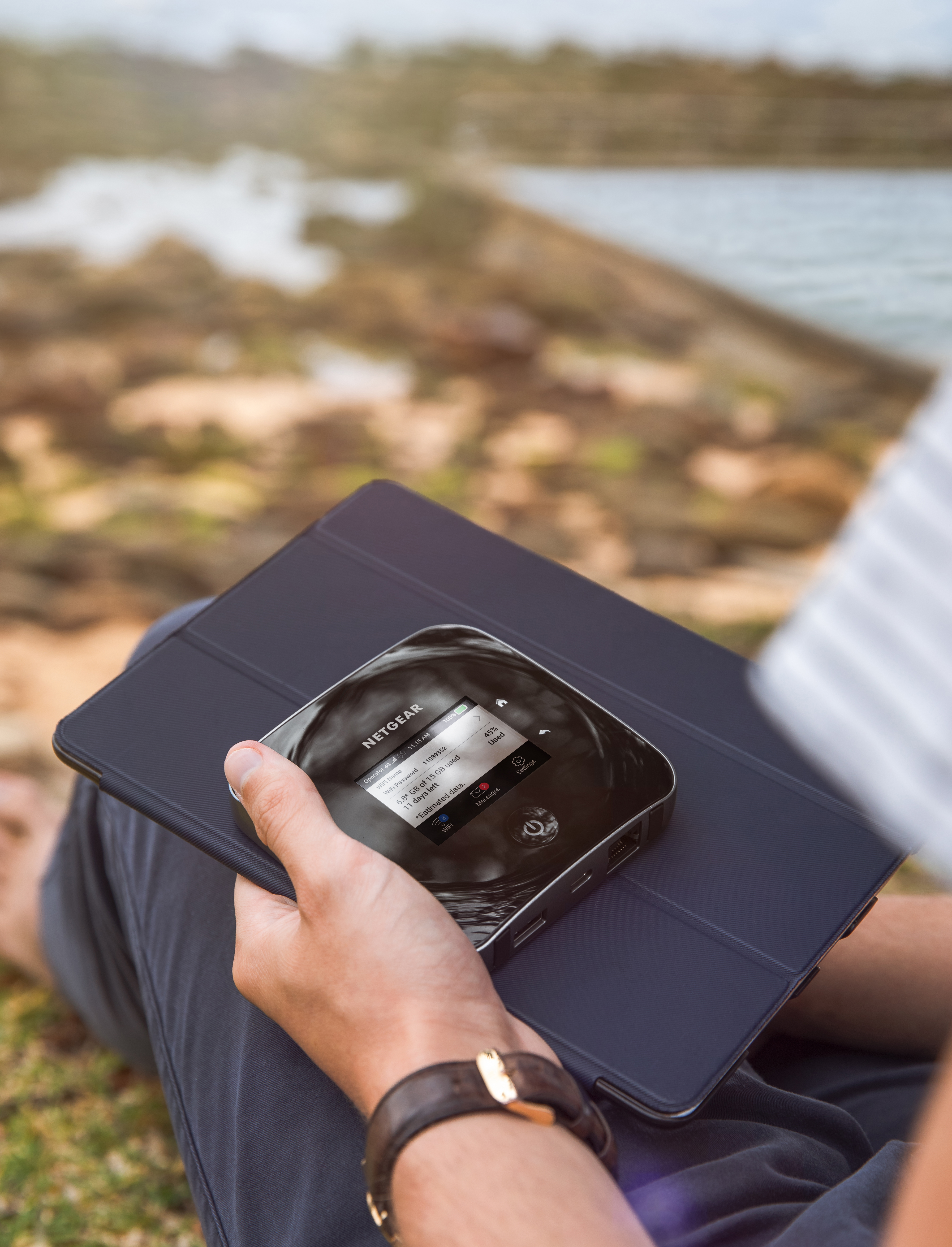 A person using a Netgear device. Sitting outside, near water. Tablet is next to the device.