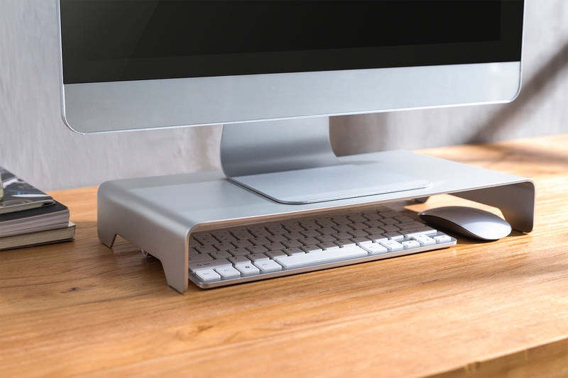 Silberner Monitorständer mit Tastatur und Maus auf einem Holzschreibtisch.