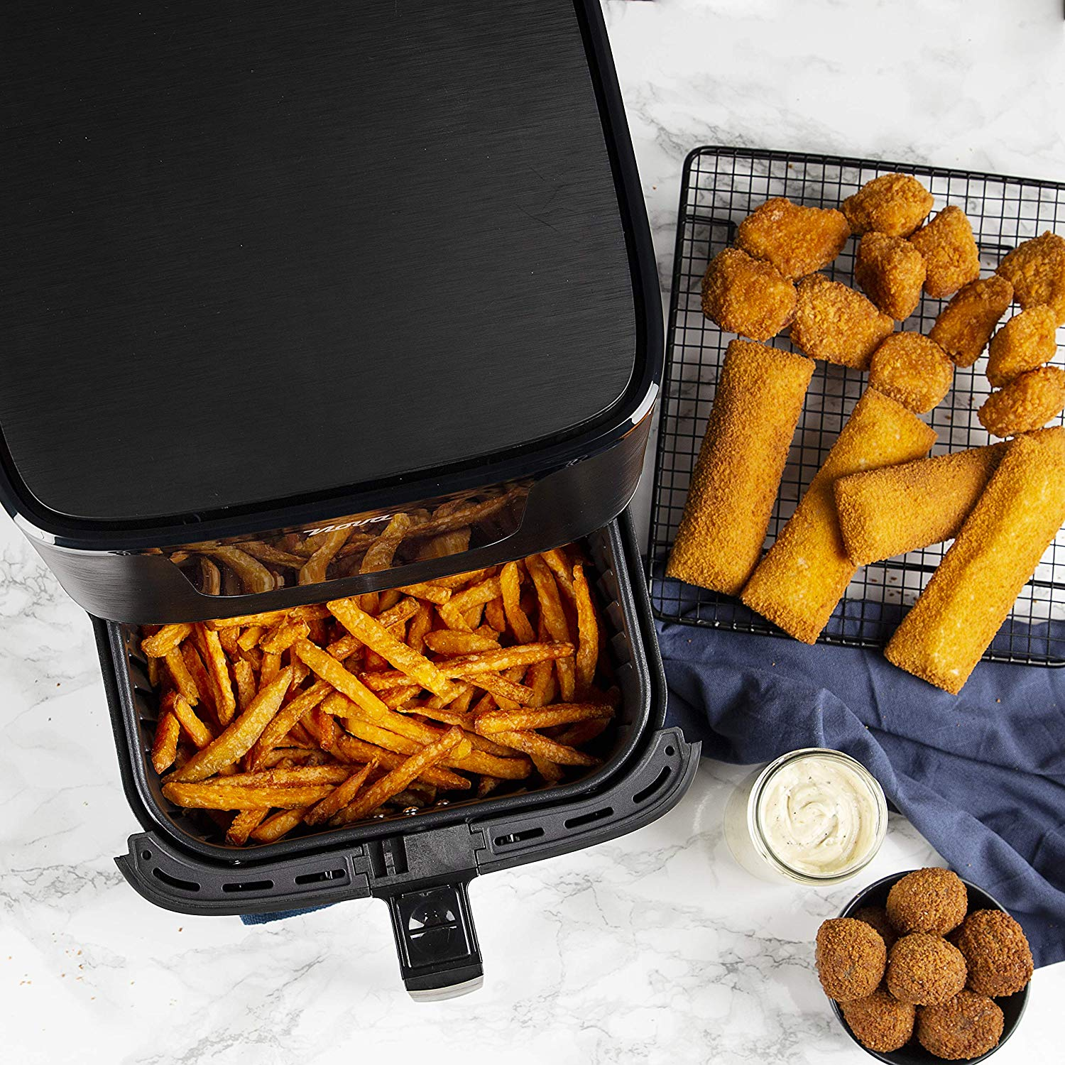 Air fryer with fries inside, with other fried foods on a rack. A small bowl of sauce is present.