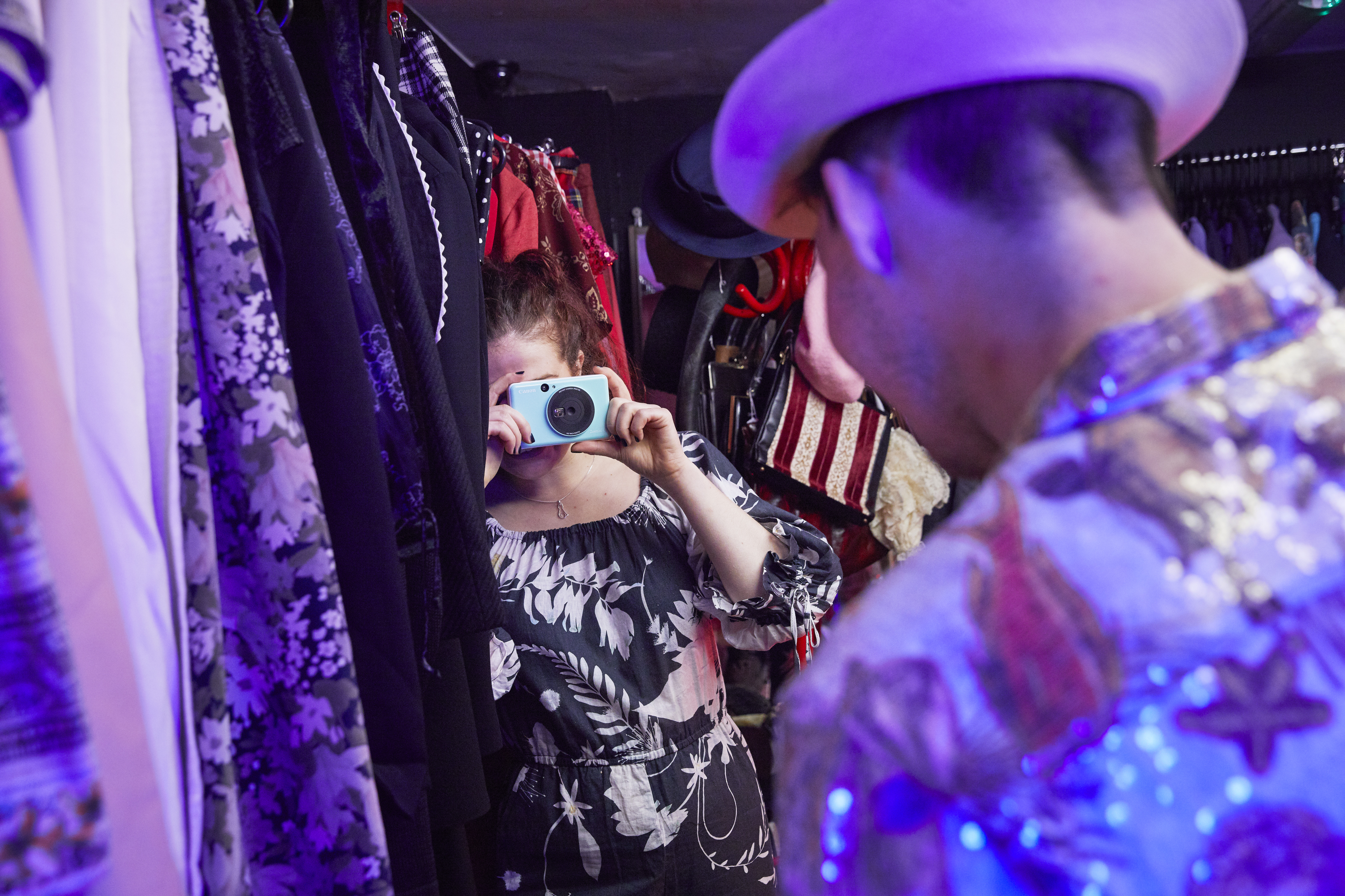 Woman taking a photo with a blue camera in a shop with clothes and a man in a hat.