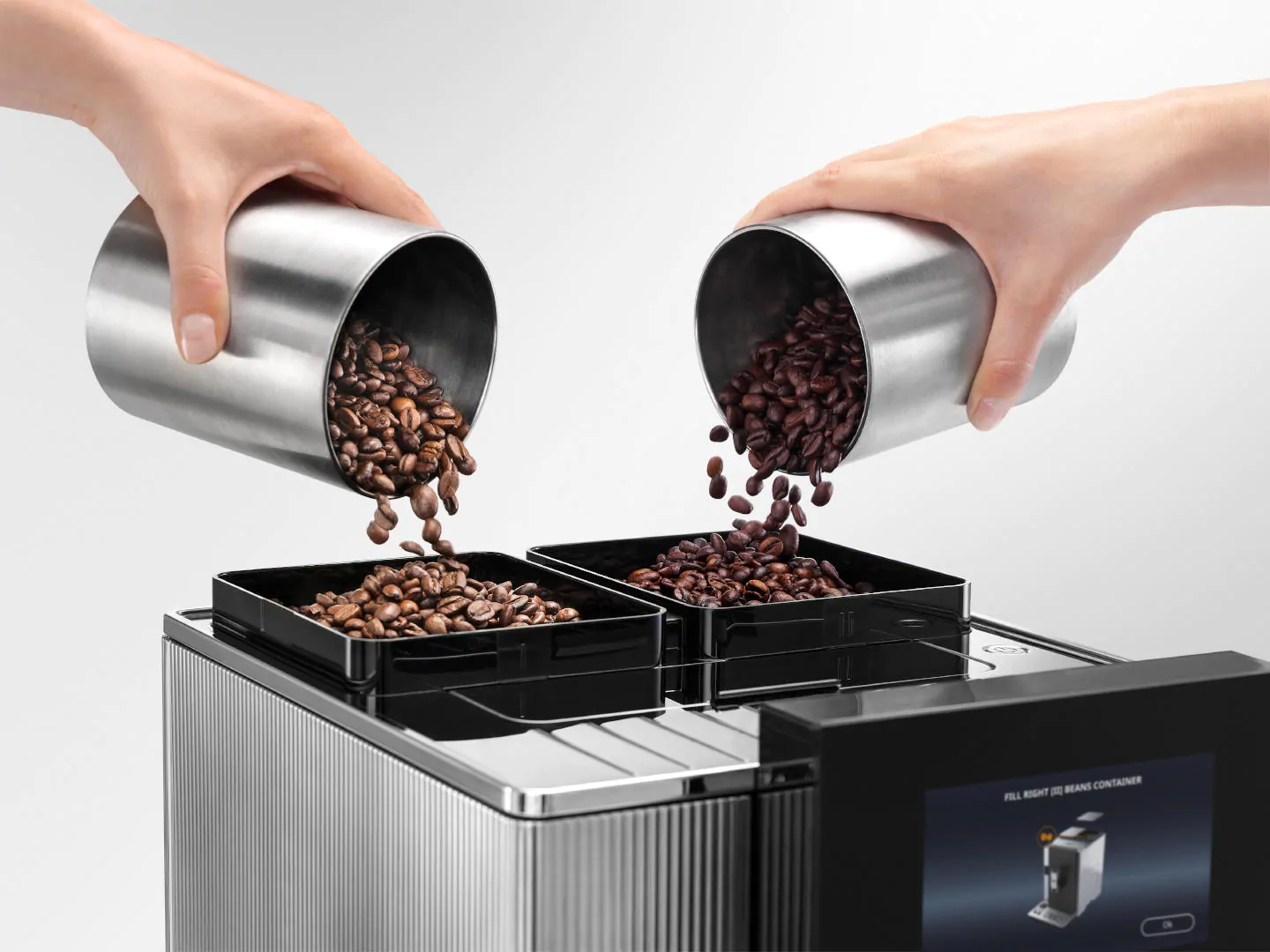 Mains versant des grains de café dans deux contenants noirs sur une machine à café. Le fond est blanc.