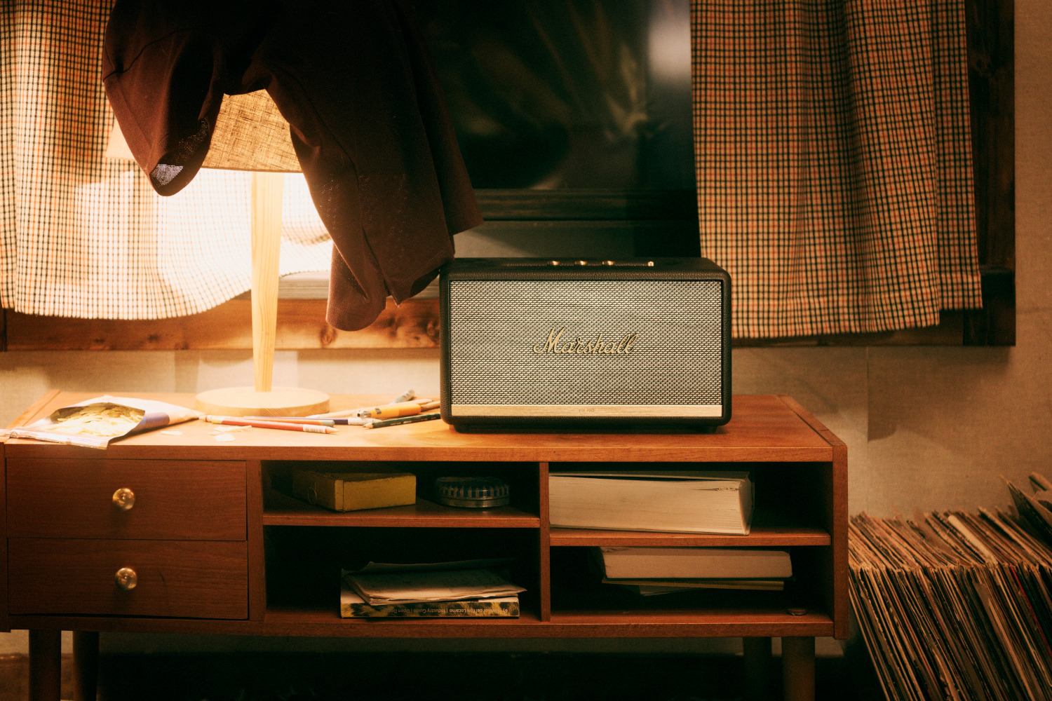 Une enceinte Marshall sur une commode en bois. Il y a une lampe et une fenêtre en arrière-plan. La scène est chaleureuse.