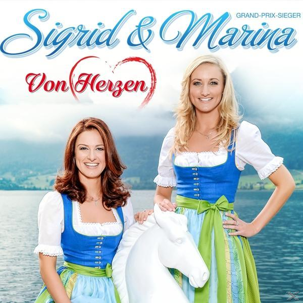 Zwei Frauen in Trachten posieren mit einer weißen Pferdestatue in der Nähe von Wasser und Bergen.