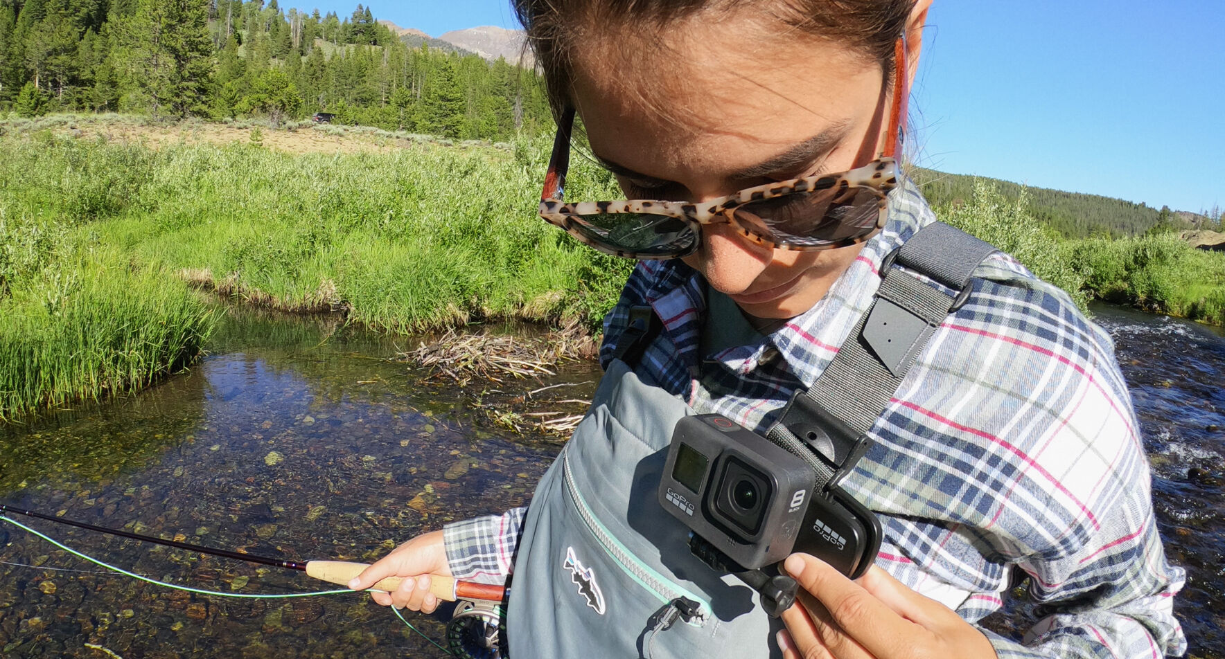 Frau fischt in einem Bach mit einer GoPro-Kamera an der Brust, trägt eine Brille und ein kariertes Hemd.
