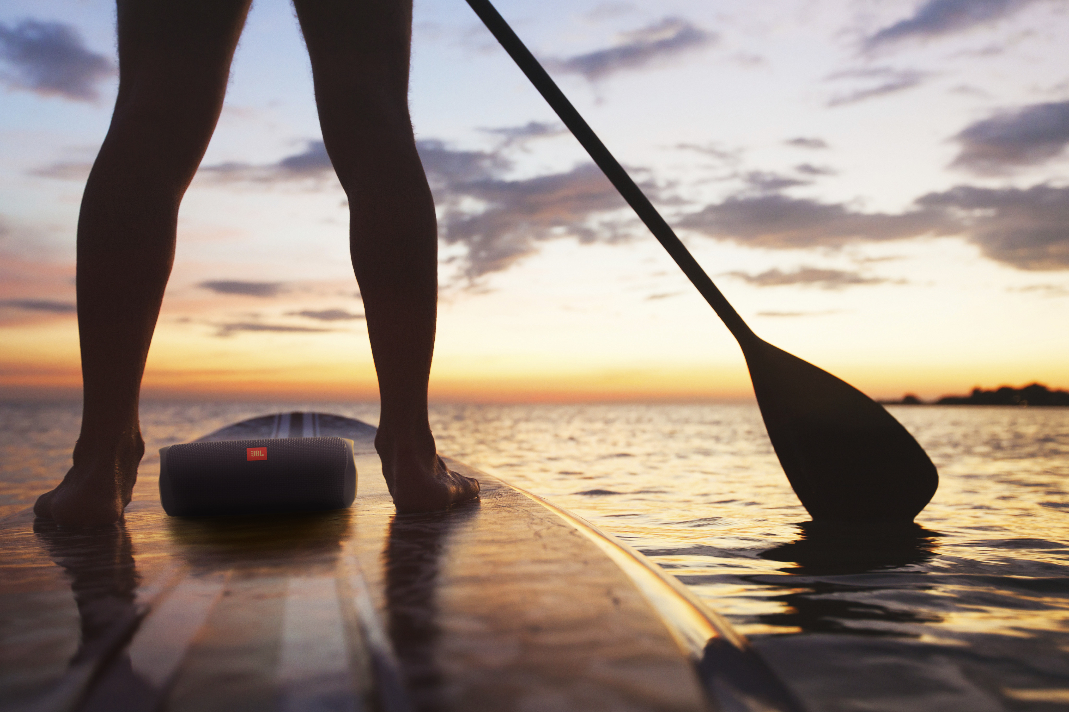 Silhouet van benen op een paddleboard. Peddel en JBL-luidspreker zijn zichtbaar. Zonsonderganglucht boven het water.