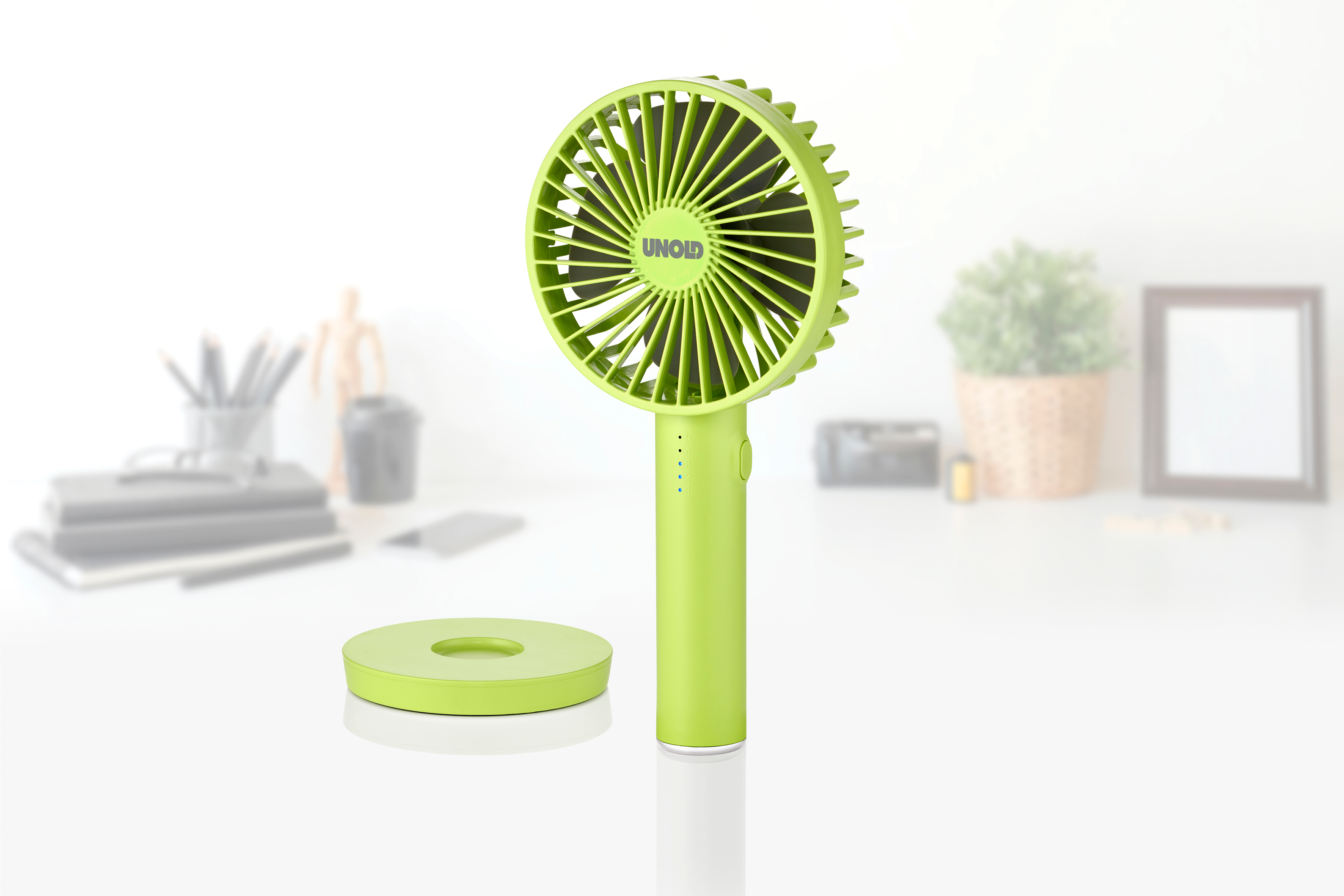 Ein grüner Handventilator steht neben einer Ladestation. Im Hintergrund sind Büroartikel.