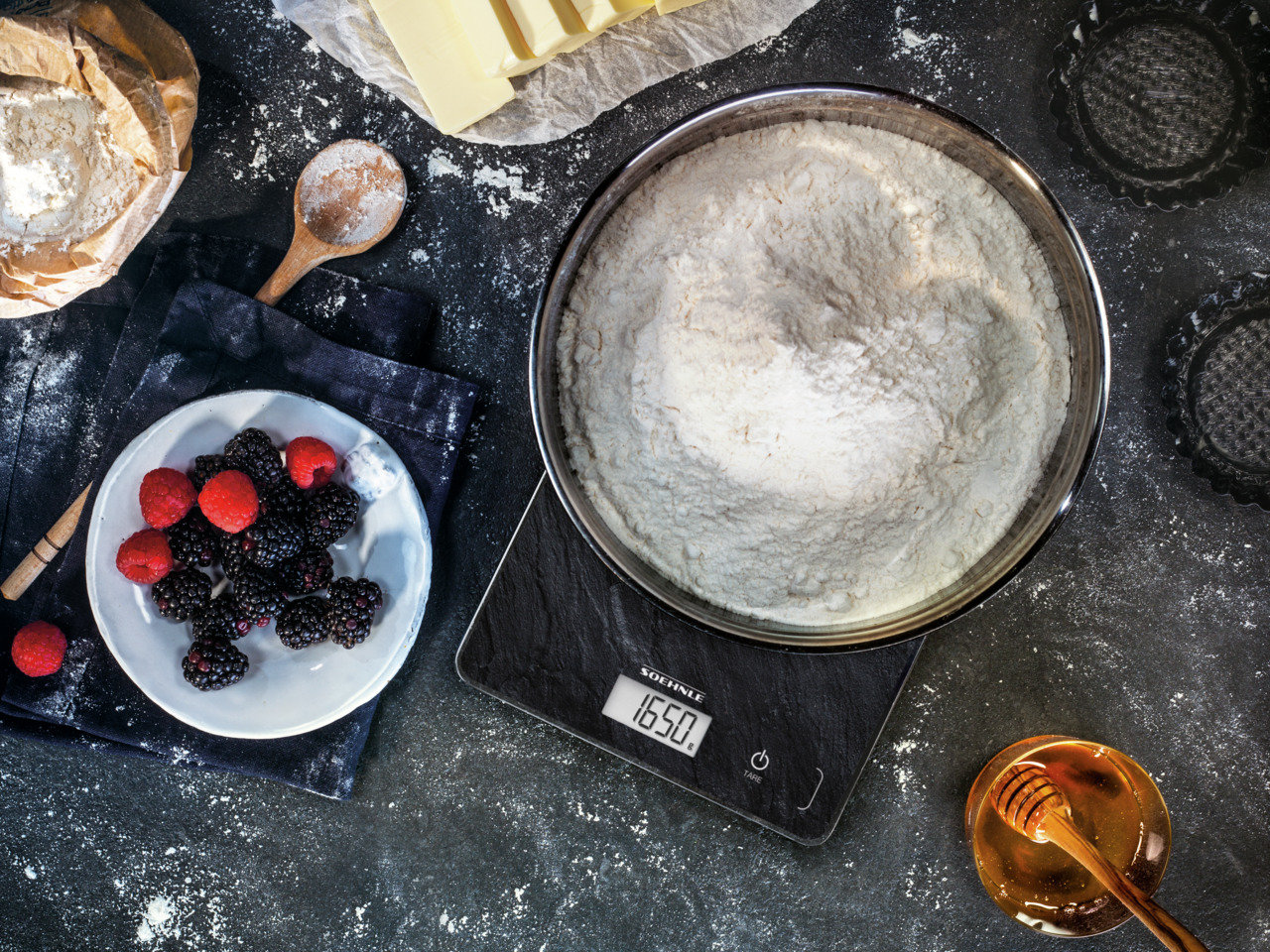 Eine Küchenszene. Mehl, Beeren, Waage, Honig, Butter und ein Holzlöffel liegen auf einer Oberfläche.