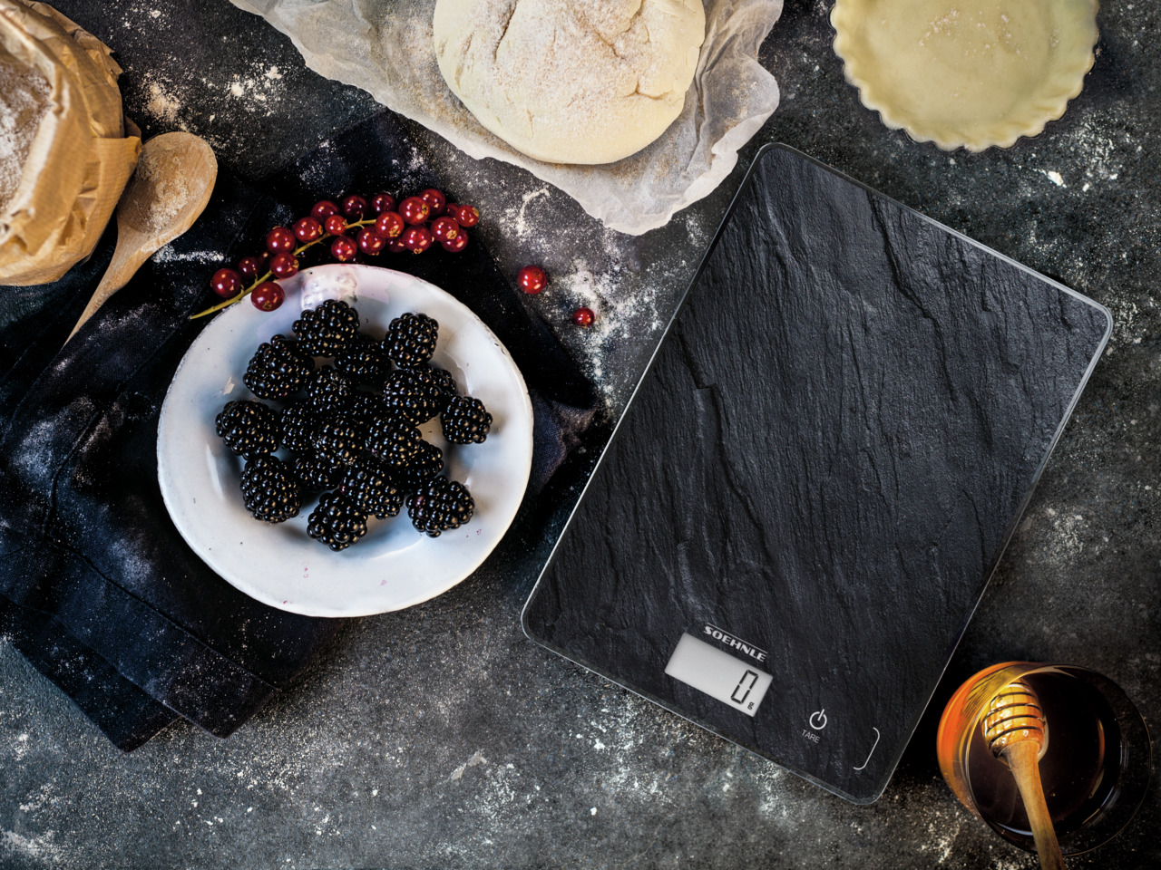 Brombeeren in einer weißen Schüssel auf dunkler Oberfläche mit Waage, Honig und Backwaren.