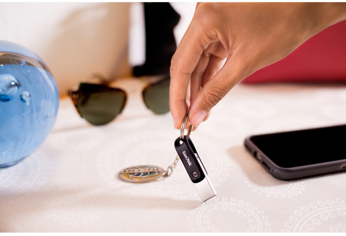 Een hand die een zwarte USB-stick met een metalen uiteinde, een sleutelhanger en een telefoon vasthoudt.