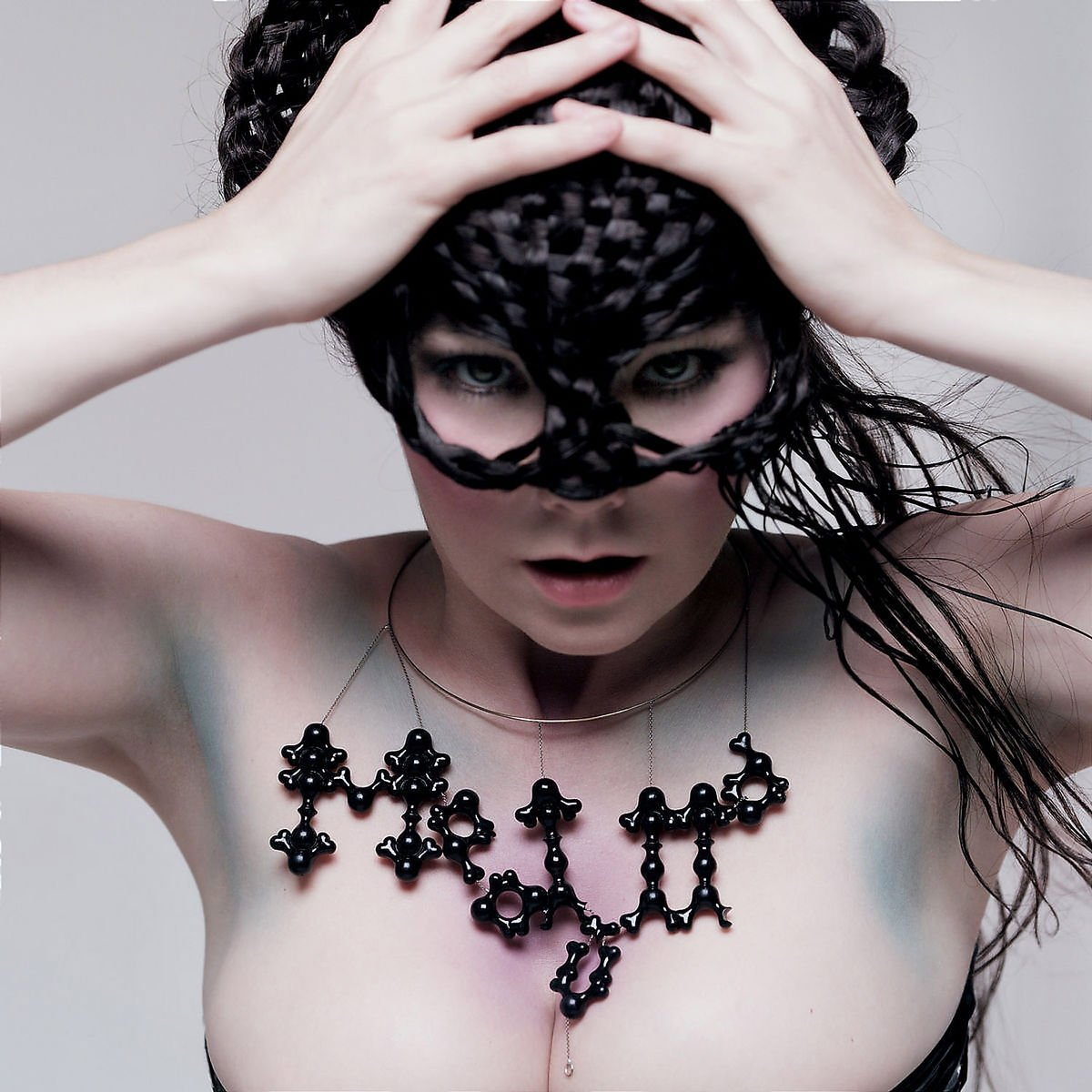Visage de Bjork avec un masque, collier et torse nu. Ses mains sur sa tête.