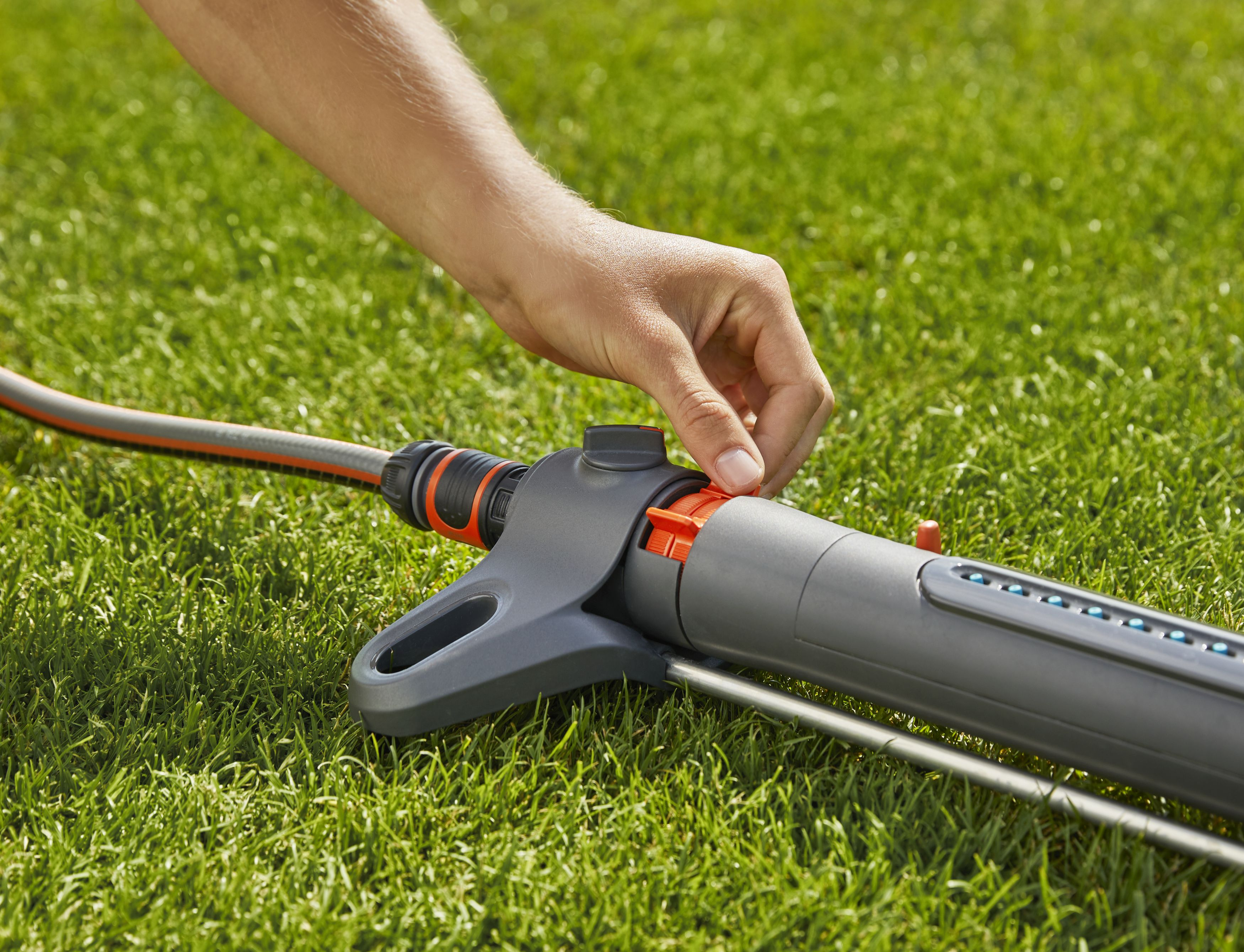 Eine Hand justiert einen grau-orangenen Gartensprenger auf grünem Gras.