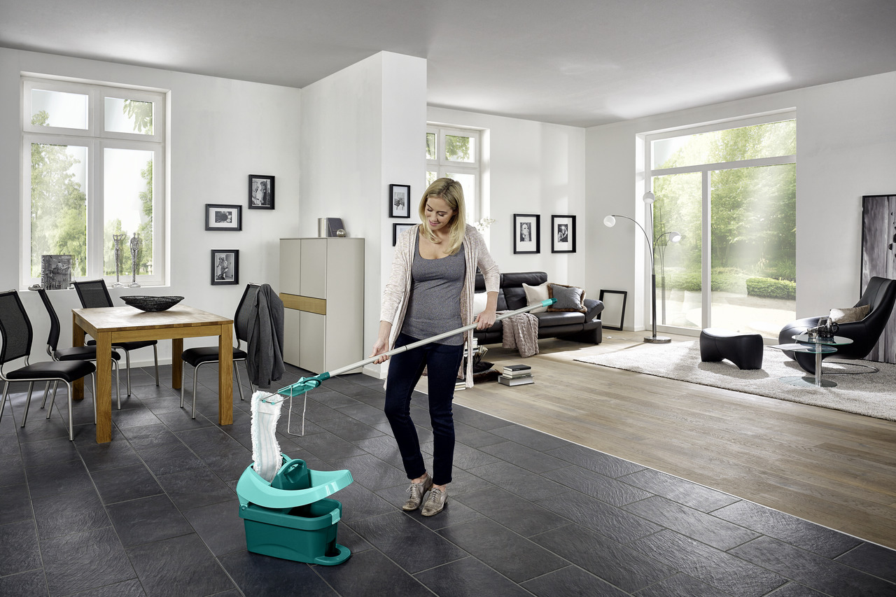 Une femme lave un sol carrelé sombre. Un seau de nettoyage est au premier plan.