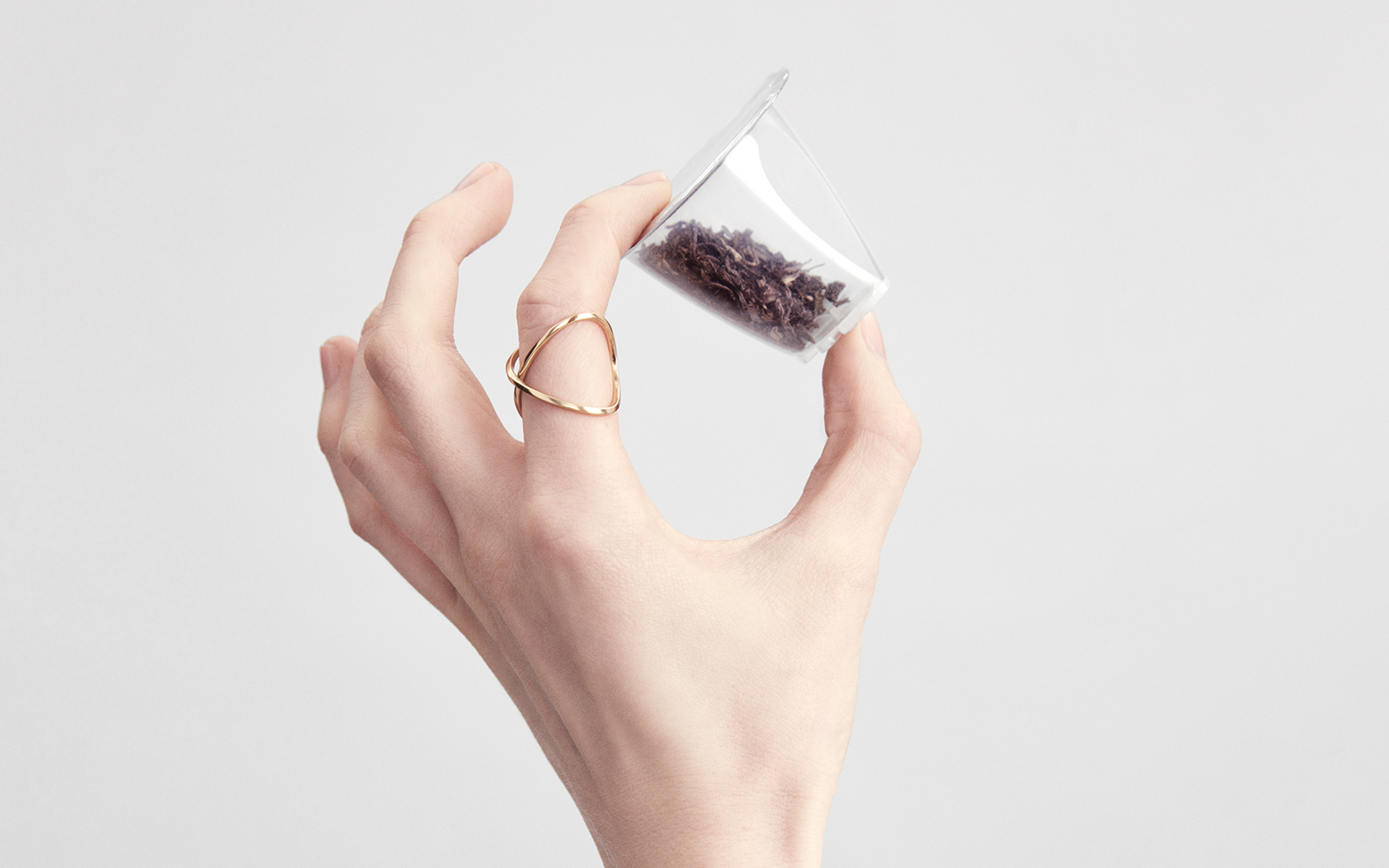 Eine Hand hält einen transparenten Plastikbehälter mit Teeblättern. Ein goldener Ring ist am Finger.