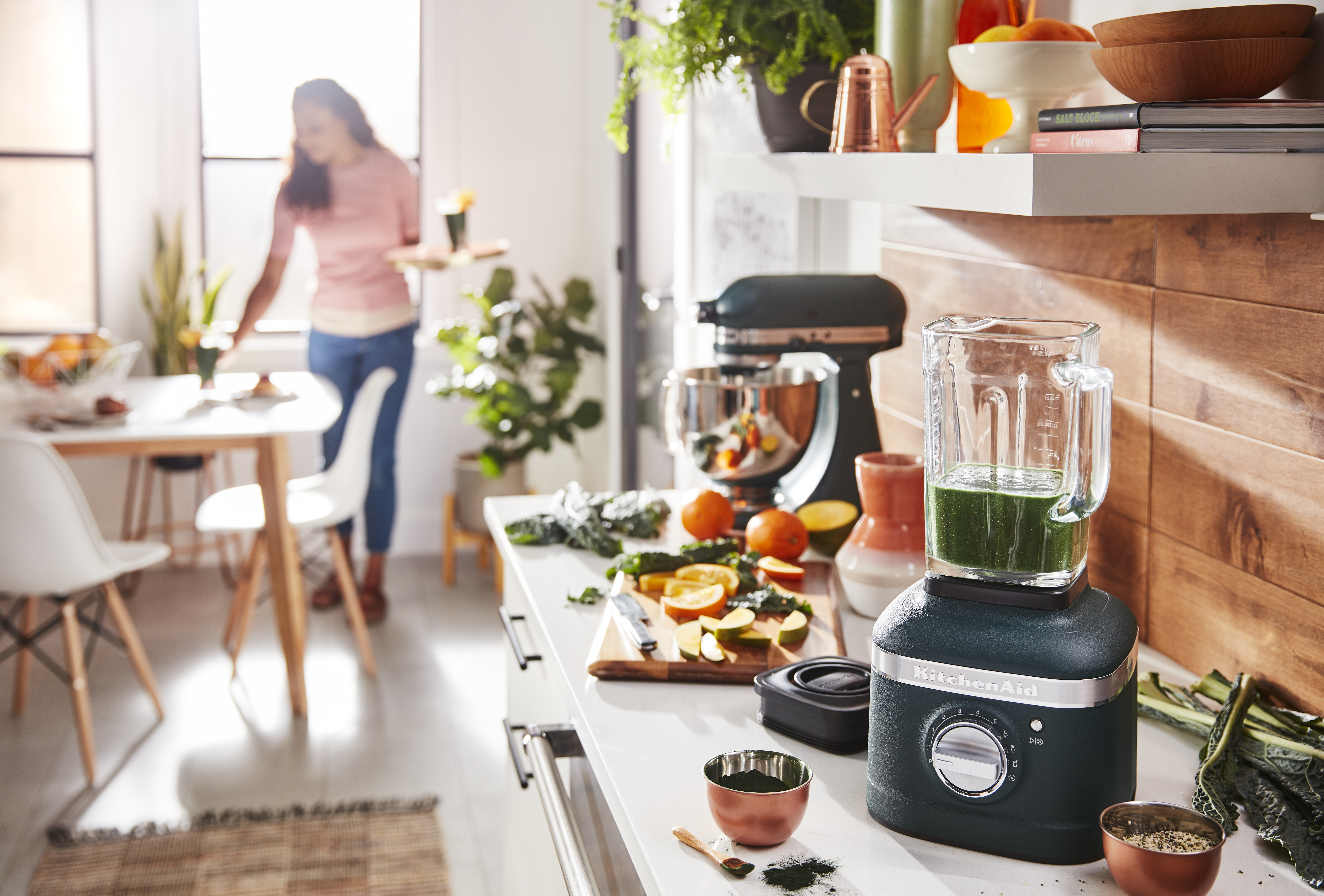 Küchenszene. Ein Mixer mit grüner Flüssigkeit. Frau im Hintergrund. Obst und Gemüse auf der Theke. KitchenAid-Mixer.