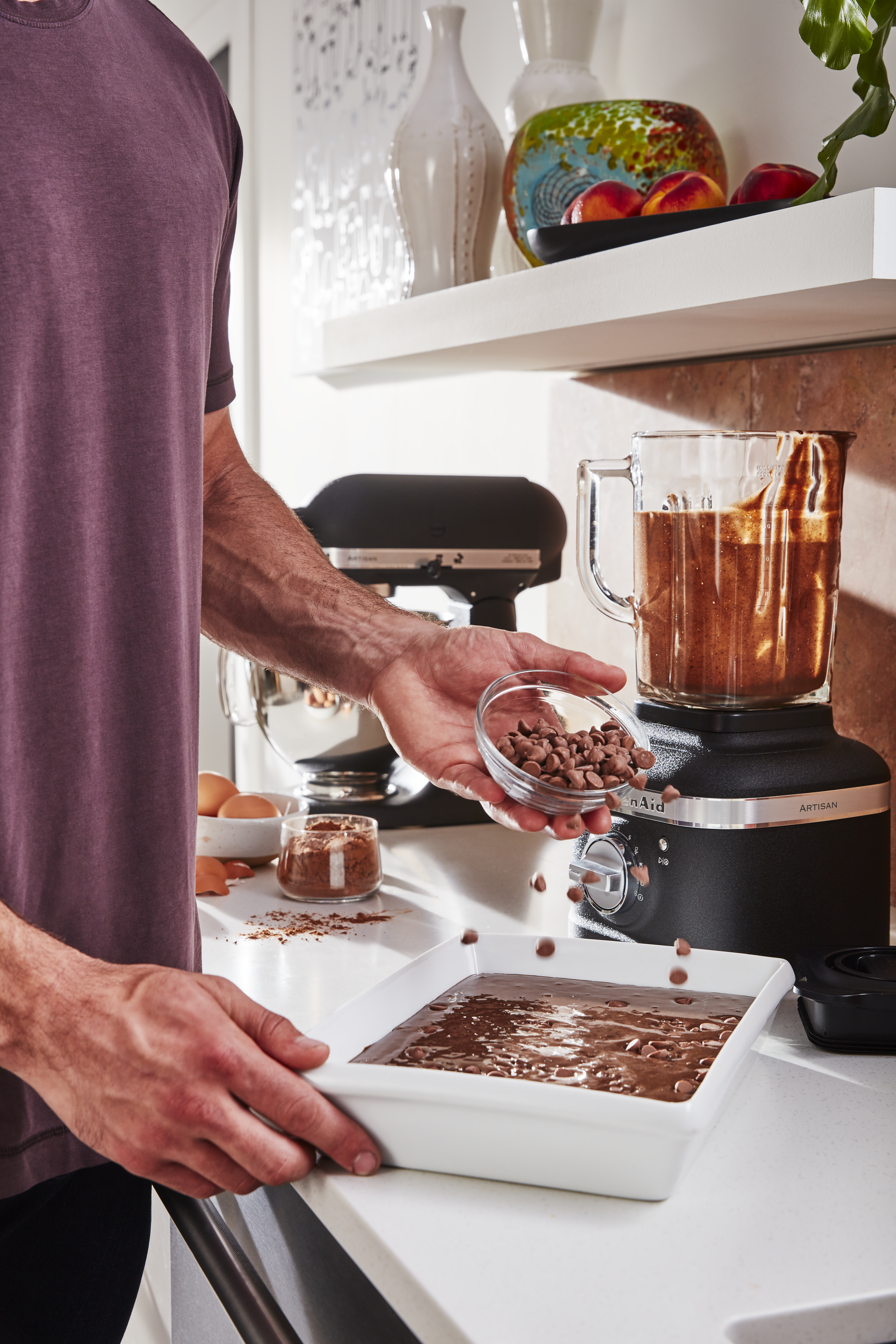 Eine Person gibt Schokoladenstückchen zu Brownie-Teig in einer weißen Schüssel. Ein Mixer mit Teig befindet sich im Hintergrund. Küchenumgebung.