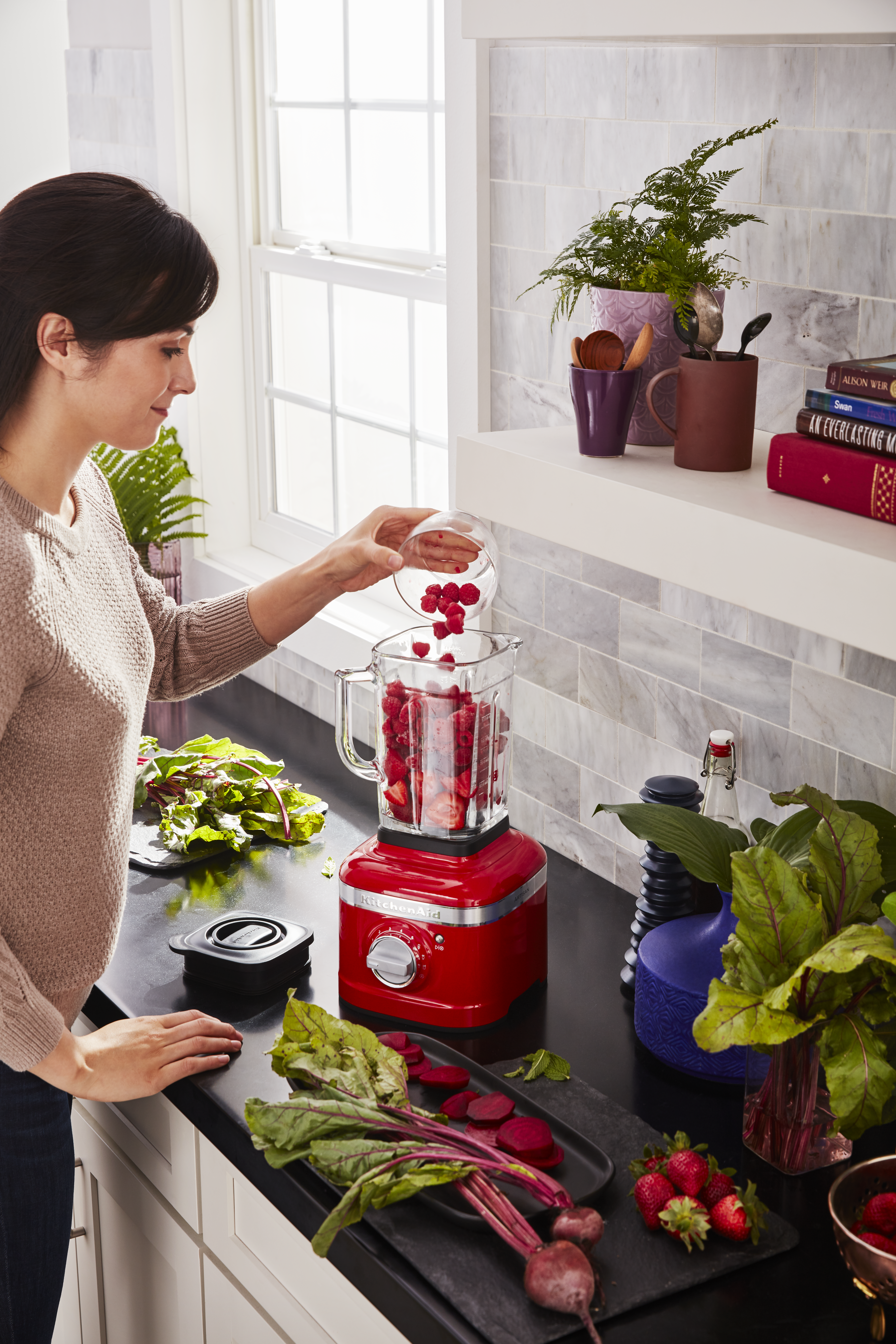 Eine Frau gibt Himbeeren in einen Mixer mit Erdbeeren. Rote Bete, Gemüse und ein roter KitchenAid-Mixer stehen auf der Theke.