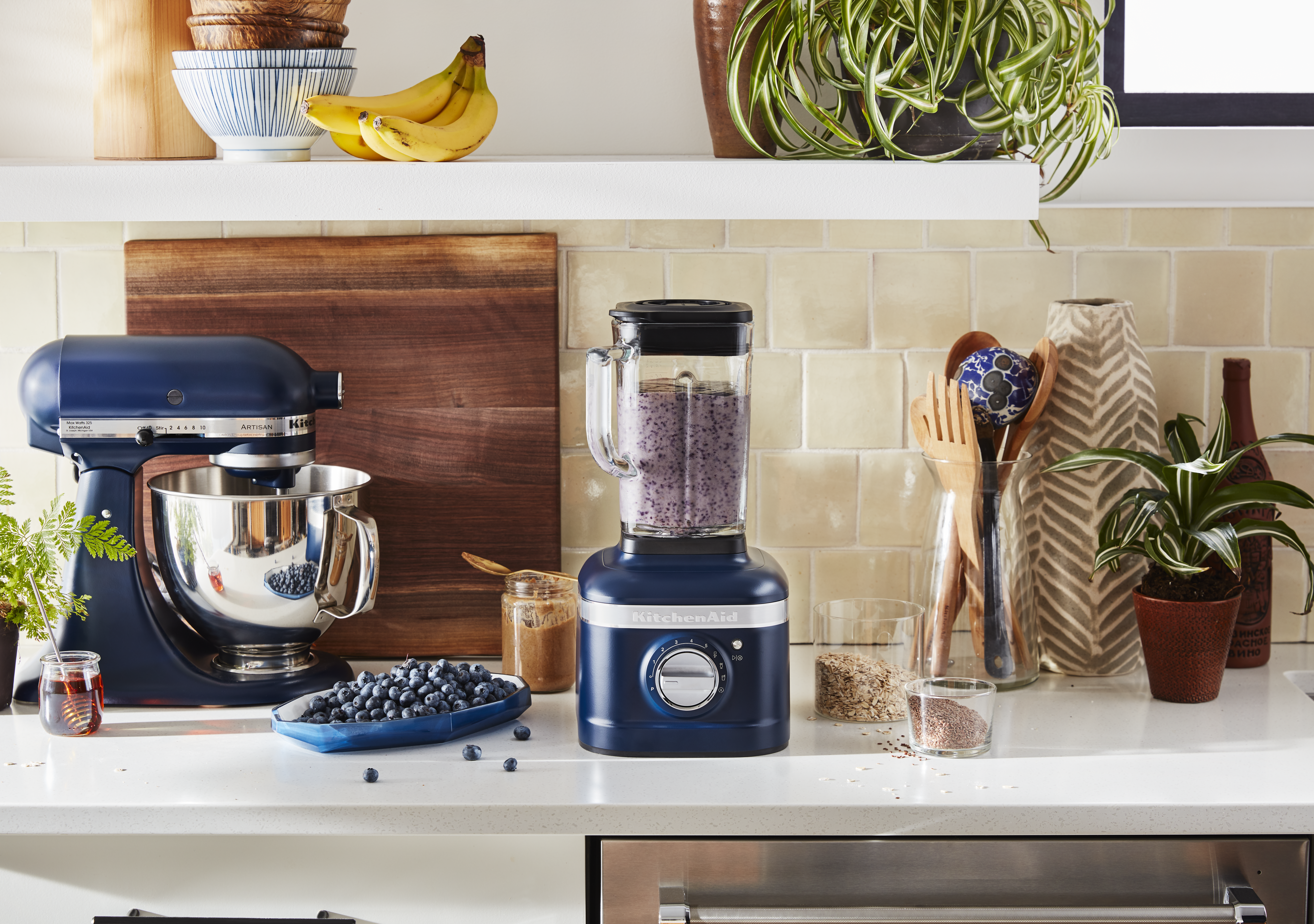Eine Küchenszene mit einem blauen Mixer, Mixer und Obst. Zutaten und Pflanzen sind auf einer weißen Arbeitsplatte und einem Regal angeordnet.