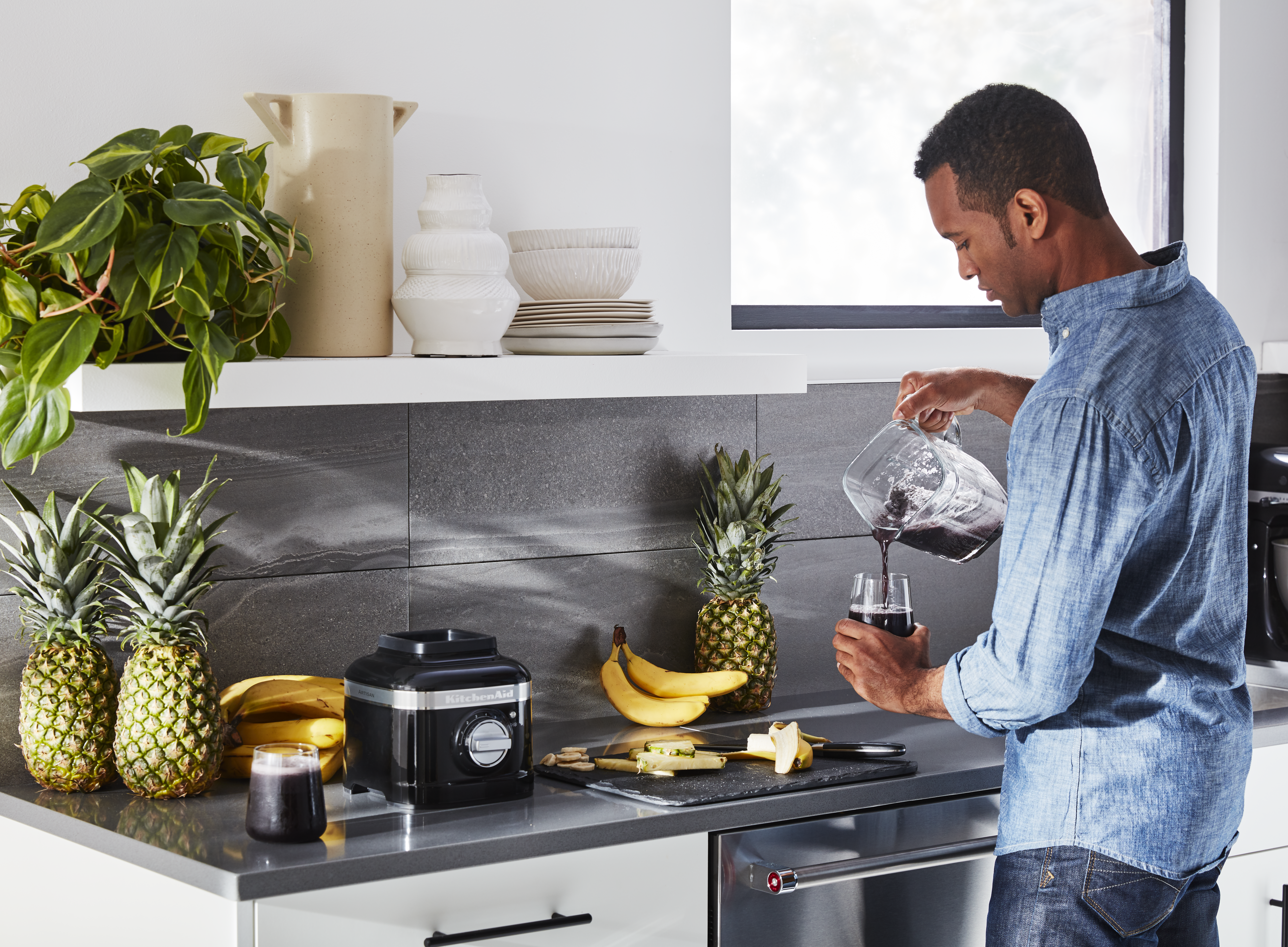 Ein Mann gießt Saft in ein Glas. Ananas, Bananen und ein Mixer stehen auf der Theke. Pflanzen und Geschirr stehen auf einem Regal.