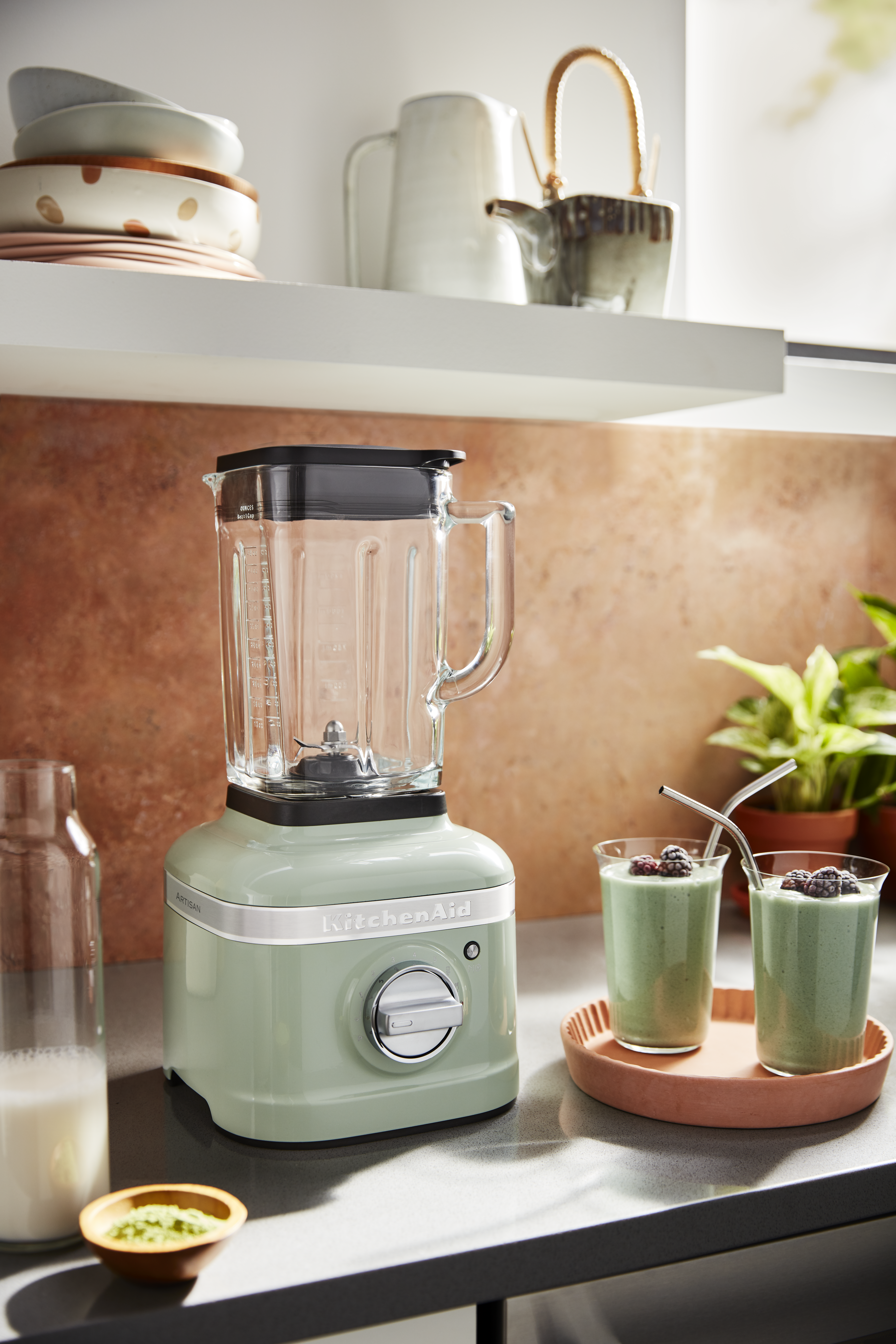 Grüner KitchenAid-Blender mit zwei Smoothies, Milch und Matcha. Regale mit Geschirr und einer Teekanne. Pflanzen und eine braune Wand.