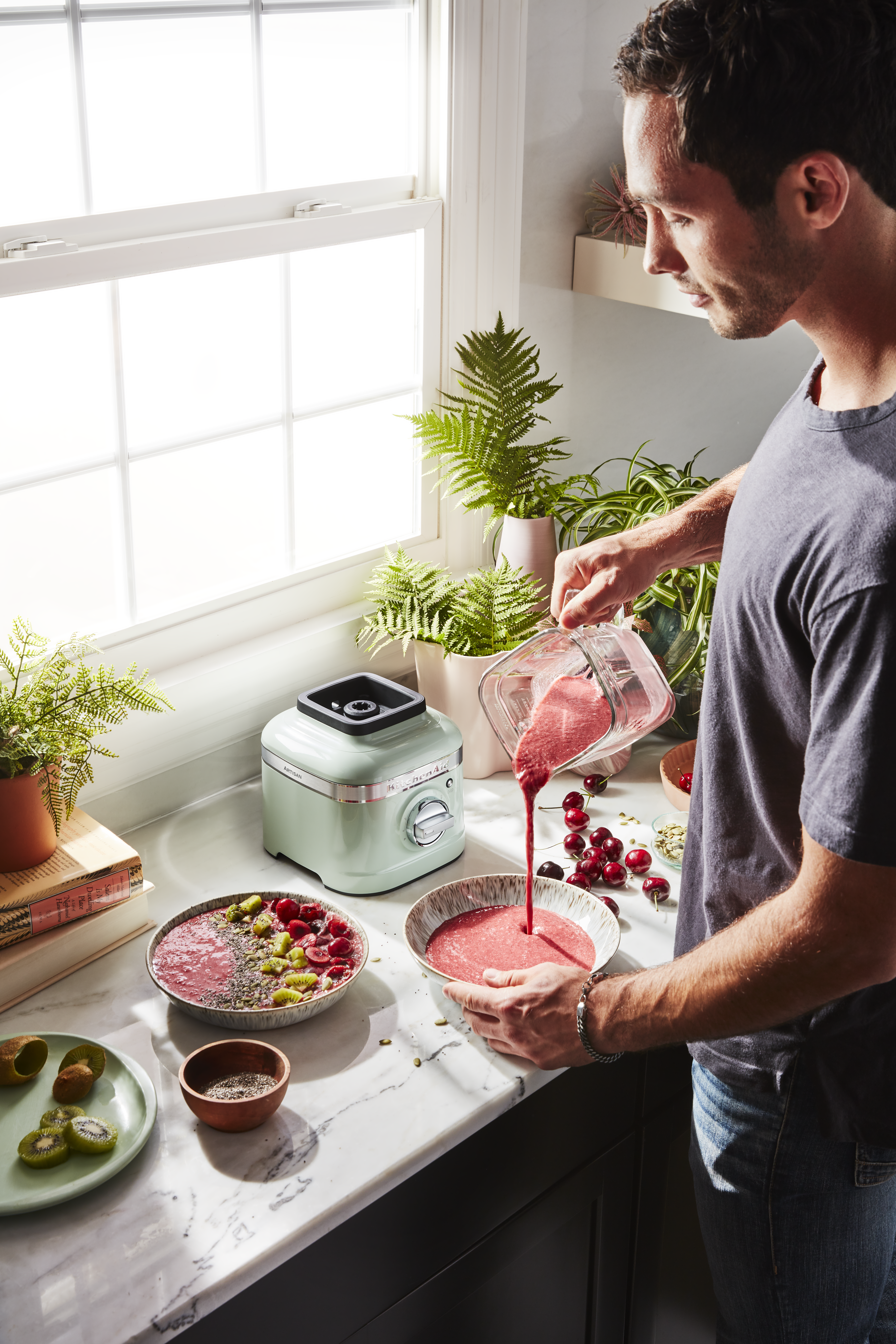 Ein Mann gießt Smoothie in eine Schüssel. Kiwi, Kirschen und Samen sind sichtbar. Ein Mixer und Pflanzen stehen auf der Theke.