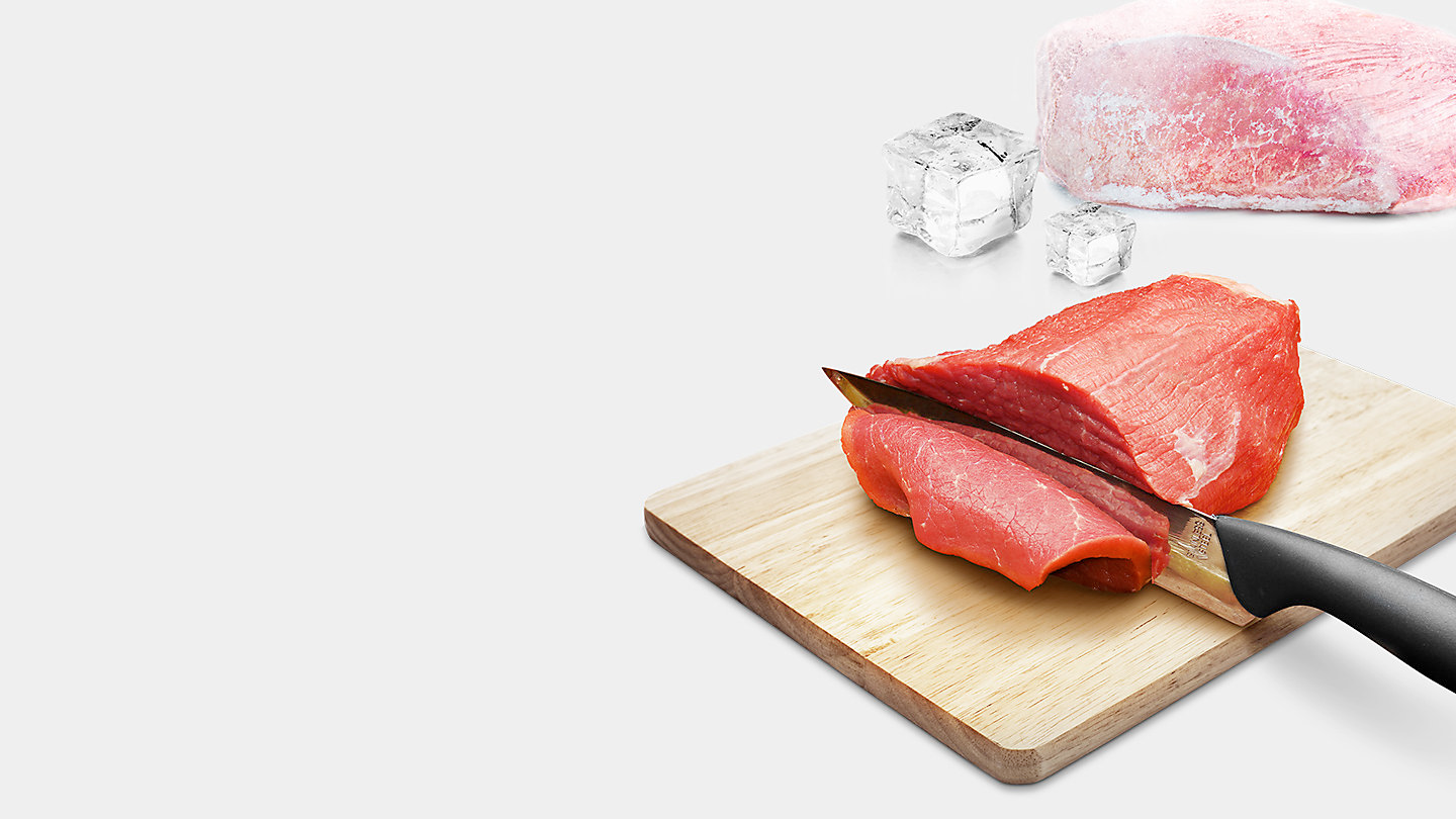 Ein Holzbrett mit einem Messer, das rohes Fleisch schneidet, mit Eis und einem großen Fleischstück im Hintergrund.