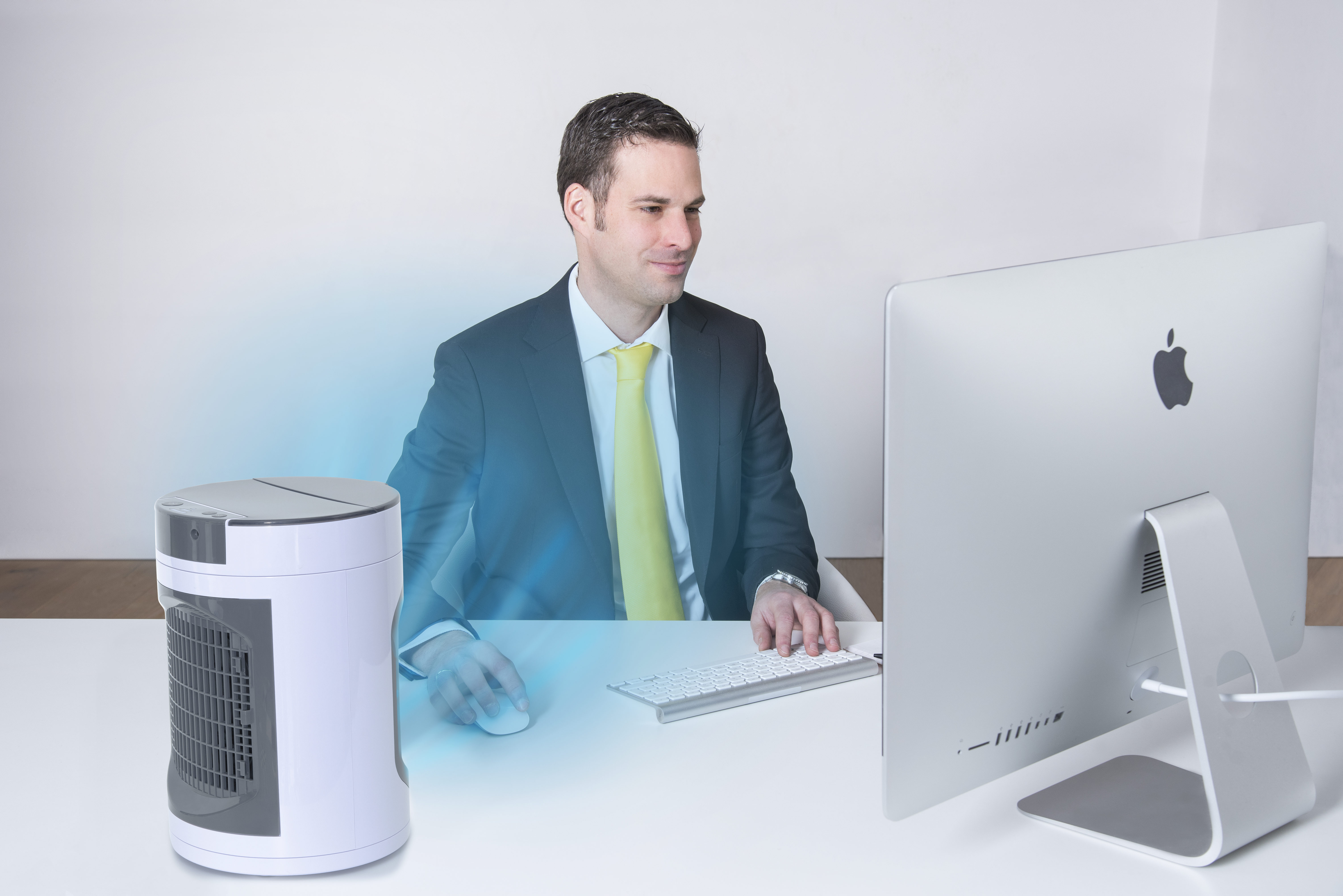 Ein Mann im Anzug sitzt an einem Schreibtisch und benutzt einen Computer. Ein kleiner Kühler steht auf dem Tisch. Eine gelbe Krawatte ist sichtbar.