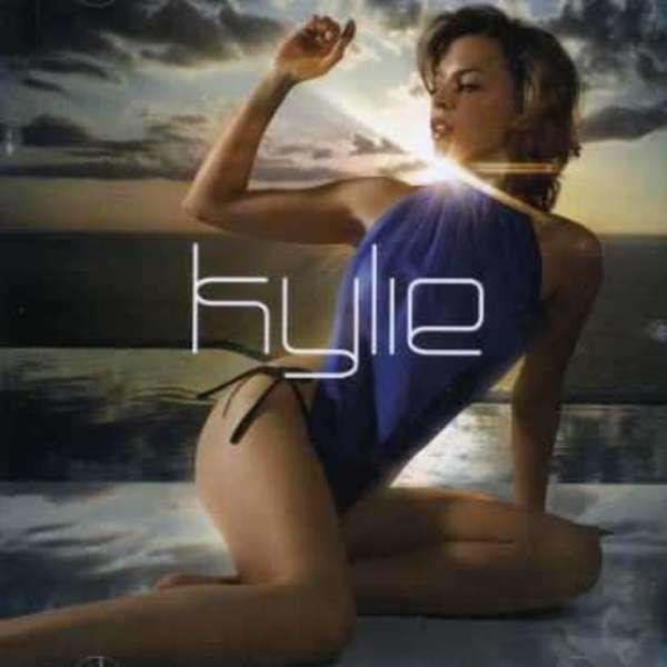 Kék fürdőruhás nő a strandon. A "Kylie" szó fehérrel van írva. A háttér egy óceán.
