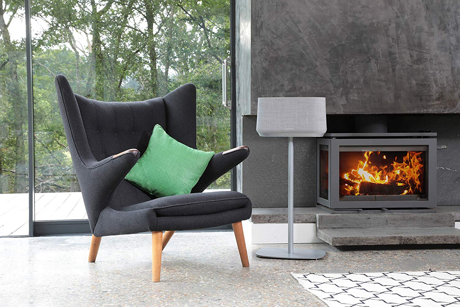 Woonkamer met fauteuil, groen kussen, speaker op standaard, open haard en groot raam.
