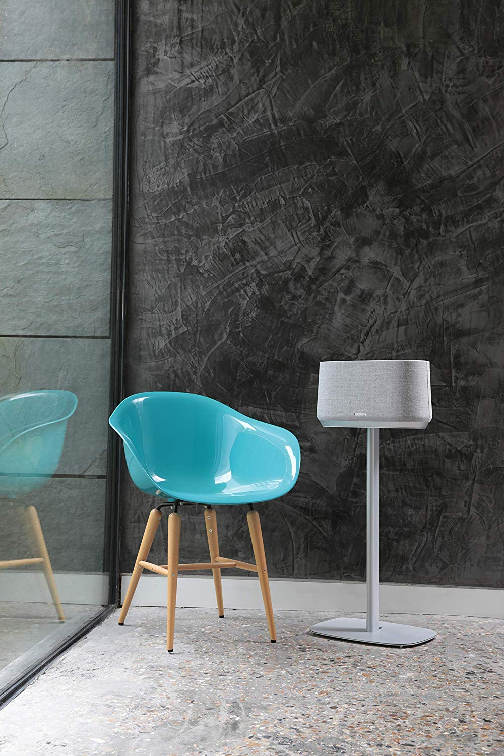 Luidspreker op zilveren standaard naast een turquoise stoel en een donkergrijze muur.