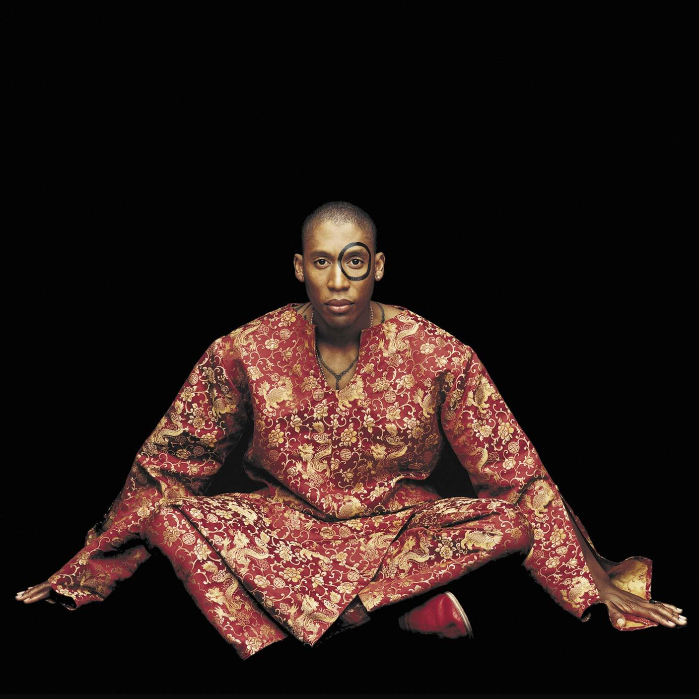 Personne en vêtements à motifs rouges et dorés, assise en tailleur.