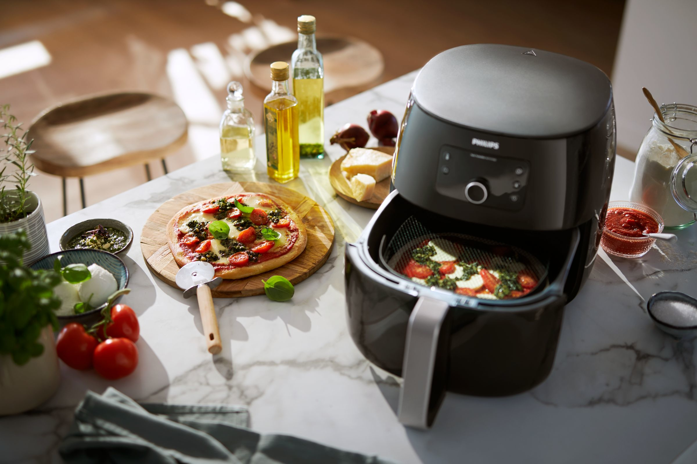 An air fryer with pizza, ingredients, and utensils on a marble countertop in a kitchen.