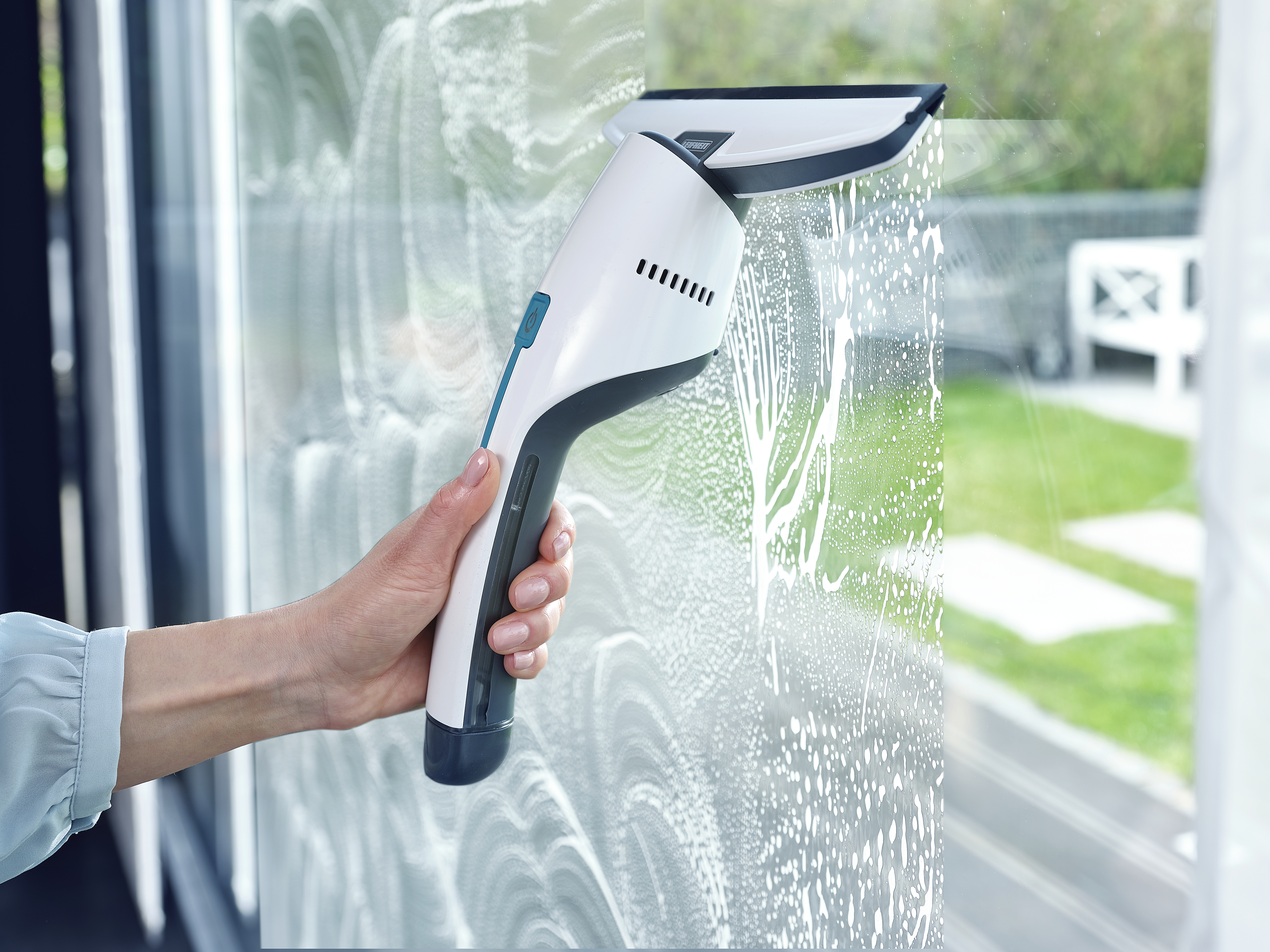 Eine Hand benutzt einen weißen und blauen Fensterreiniger, um ein Fenster mit Seifenwasser zu reinigen.