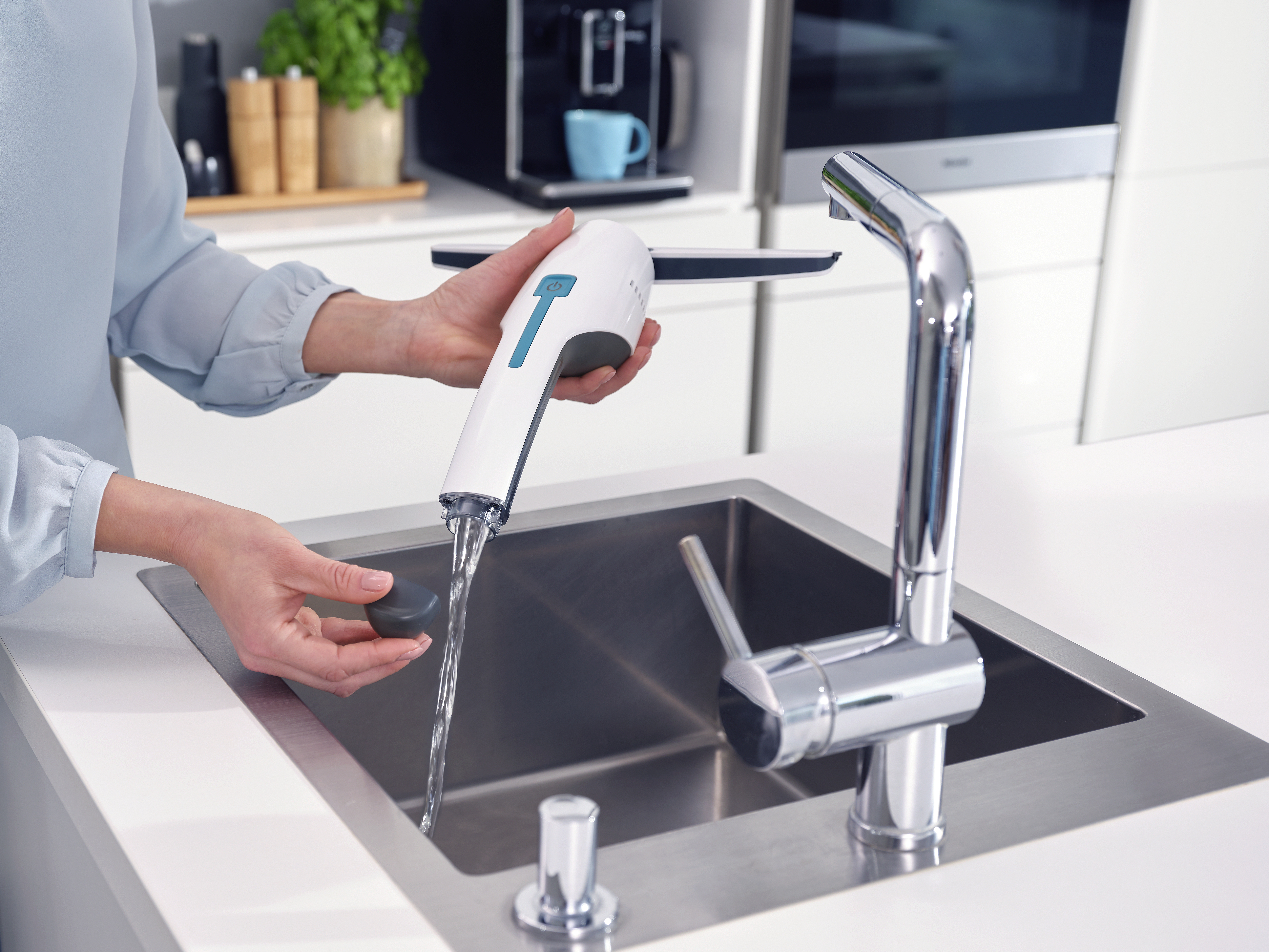Eine Frau hält ein weiß-blaues Reinigungswerkzeug über einem Edelstahlbecken mit fließendem Wasser.