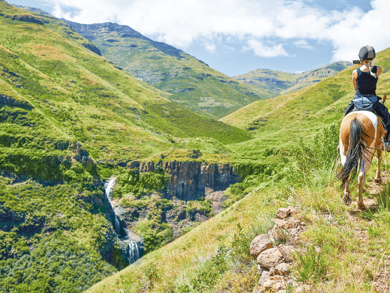Persona a cavallo su un sentiero di montagna, con vista panoramica su montagne verdi e una cascata.