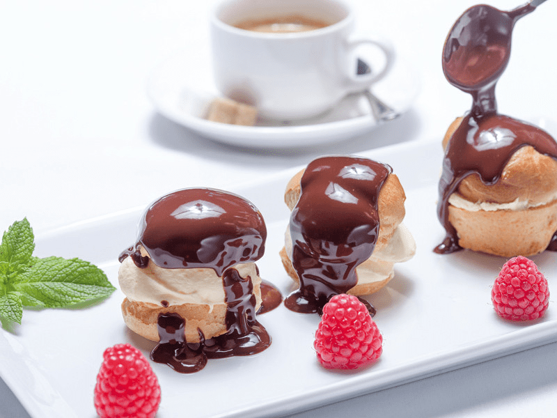 Bigné ricoperti di cioccolato su un piatto, con lamponi e una tazza di caffè.