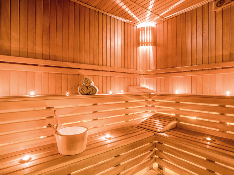 Interno di una sauna in legno. Panche, secchio di legno e asciugamani sono visibili. La scena è illuminata da piccole luci.