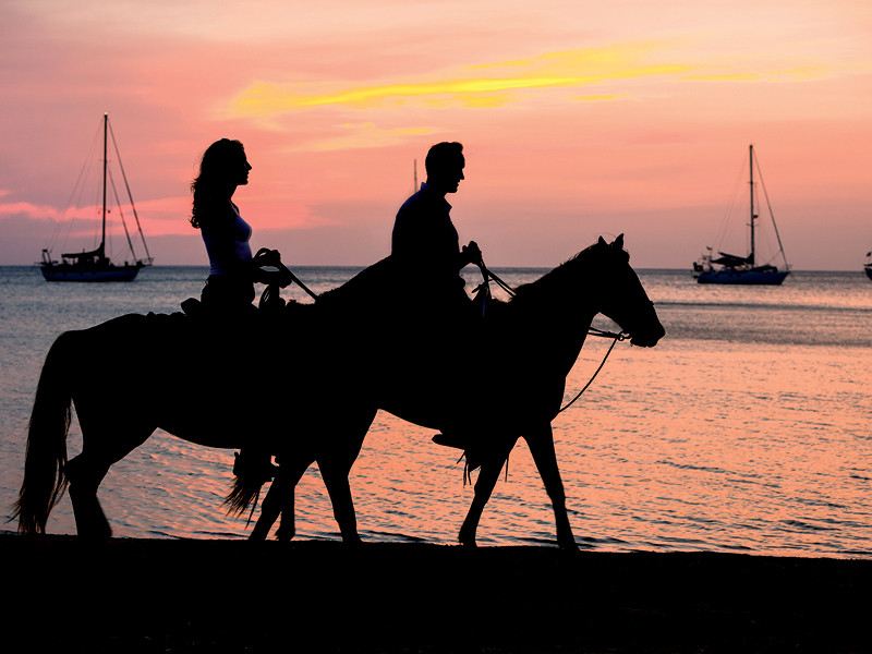 Paar reitet Pferde am Strand bei Sonnenuntergang, Segelboote im Hintergrund.