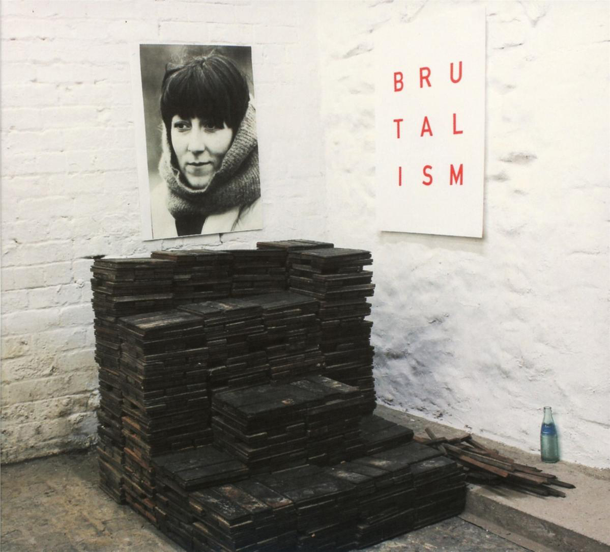 Ein Raum mit einem Stapel Holzblöcke, einem Porträt und einem Schild mit der Aufschrift „BRUTALISM“. Auf dem Boden steht eine Flasche.