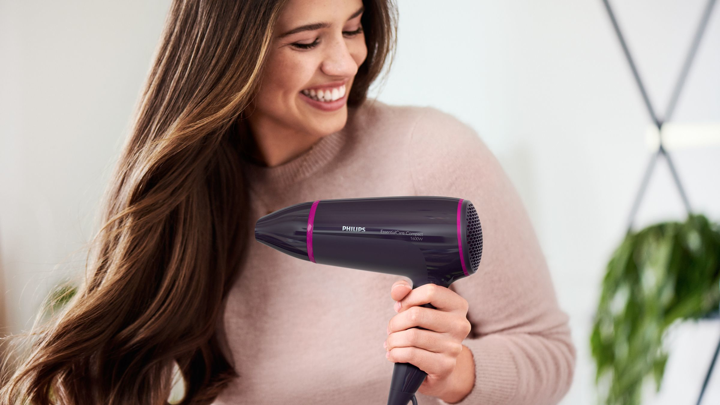 Frau mit langen, braunen Haaren lächelt, hält einen dunkelvioletten Philips Haartrockner. Grüne Pflanze im Hintergrund.