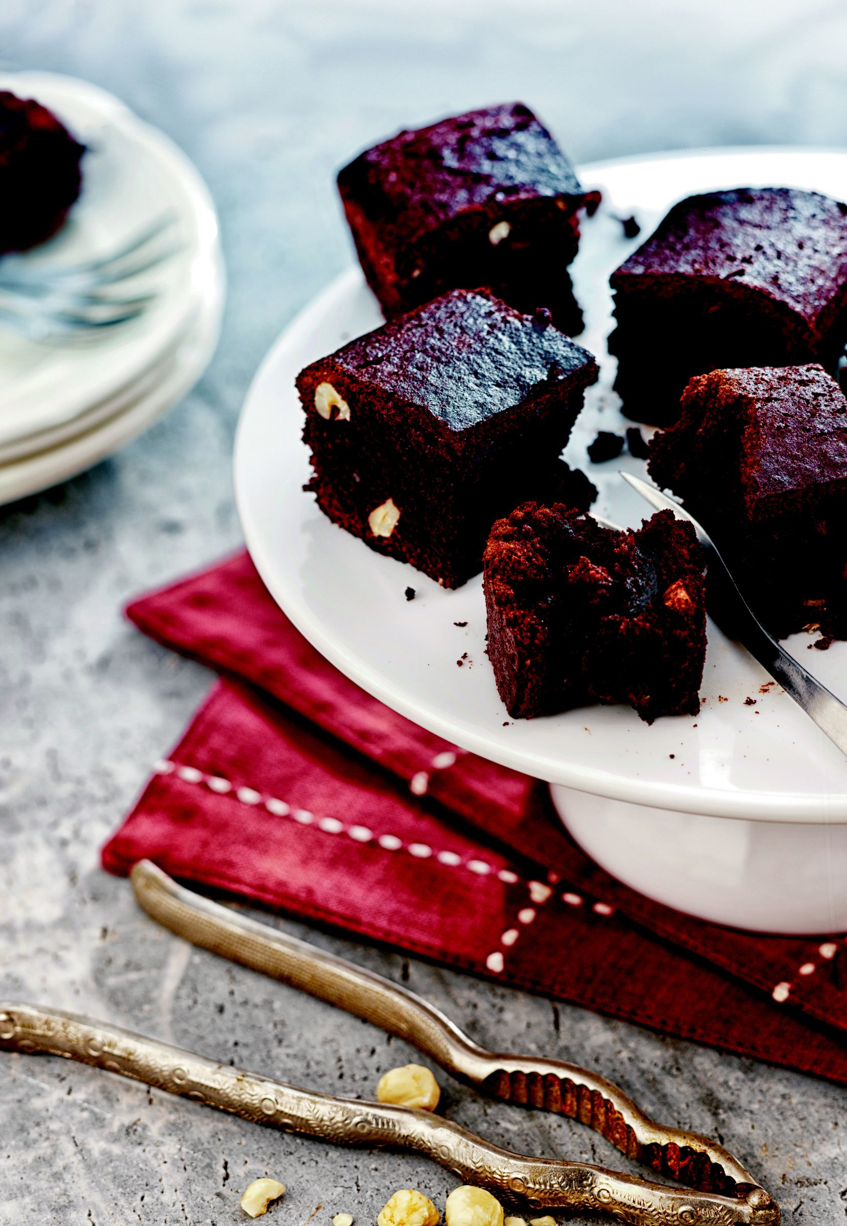 Nahaufnahme von Brownies auf einem weißen Kuchenständer. Außerdem eine Gabel, Nussknacker und Haselnüsse.