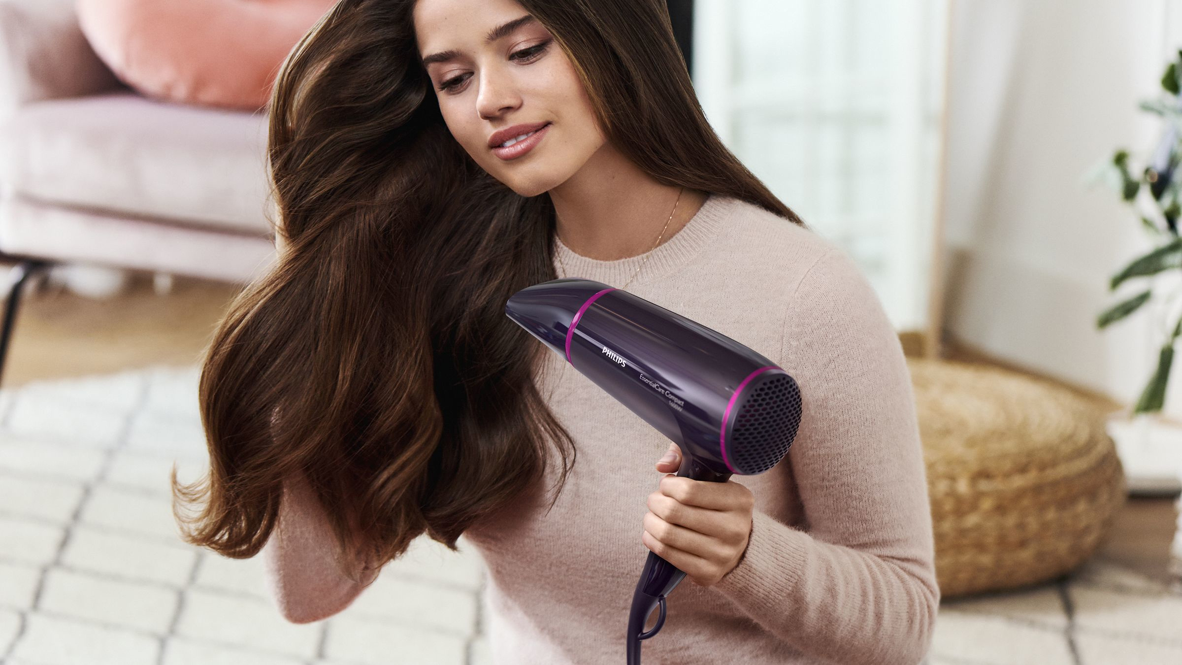 Frau trocknet ihre langen, braunen Haare mit einem dunkelvioletten Haartrockner. Sie ist in einem Raum.